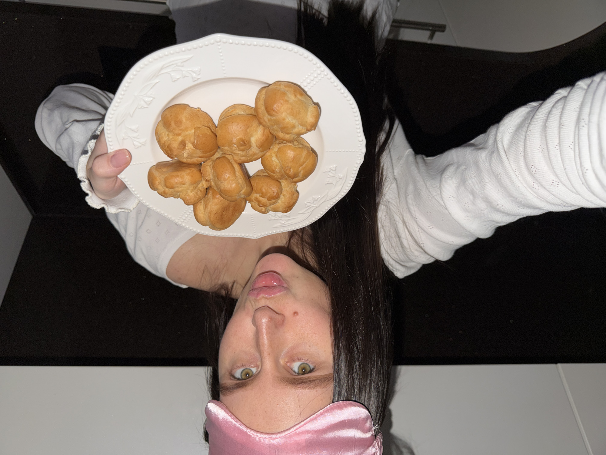 A woman wearing a pink satin sleep mask is holding up a white plate with a stack of profiteroles. The woman is wearing a white long-sleeved shirt with ruffled cuffs. Her long dark hair is loose. She has green eyes. The background appears to be a kitchen area with black countertops and white cabinets. The location is Khimki, Russia.