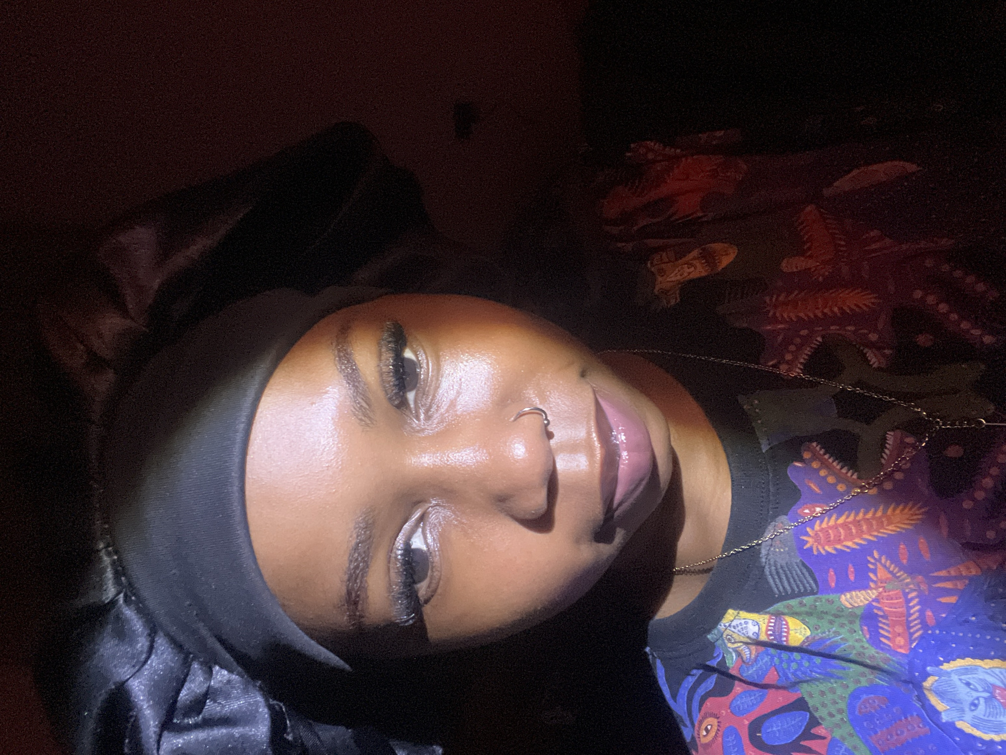 In a dimly lit interior, a young Black woman with a nose ring and dark hair covered by a black satin bonnet is captured in a close-up, frontal portrait. She is wearing a colorful, patterned t-shirt. The lighting creates a dramatic effect, casting shadows across her face and highlighting her features. The scene does not provide any visual cues to determine its specific location within Rafin Par, Nigeria.