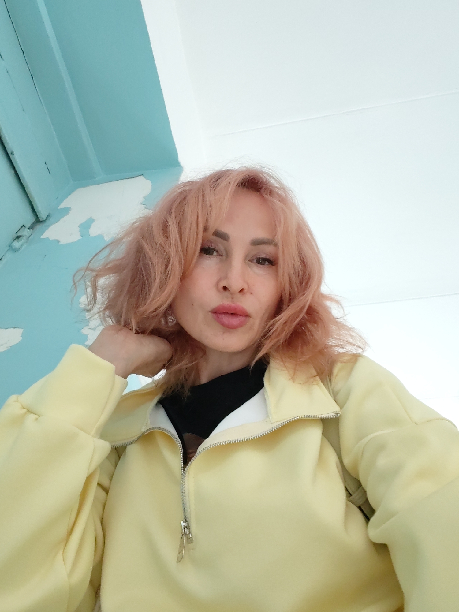 A woman with pink, wavy hair and fair skin is shown from the chest up, looking directly at the camera. She is wearing a light yellow zip-up jacket over a black garment. Her right hand is resting on her chin, and her mouth is slightly pursed. The background consists of a light blue wall with peeling white paint on the left side and a white ceiling or wall on the right. The lighting appears to be natural, coming from above and to the right.