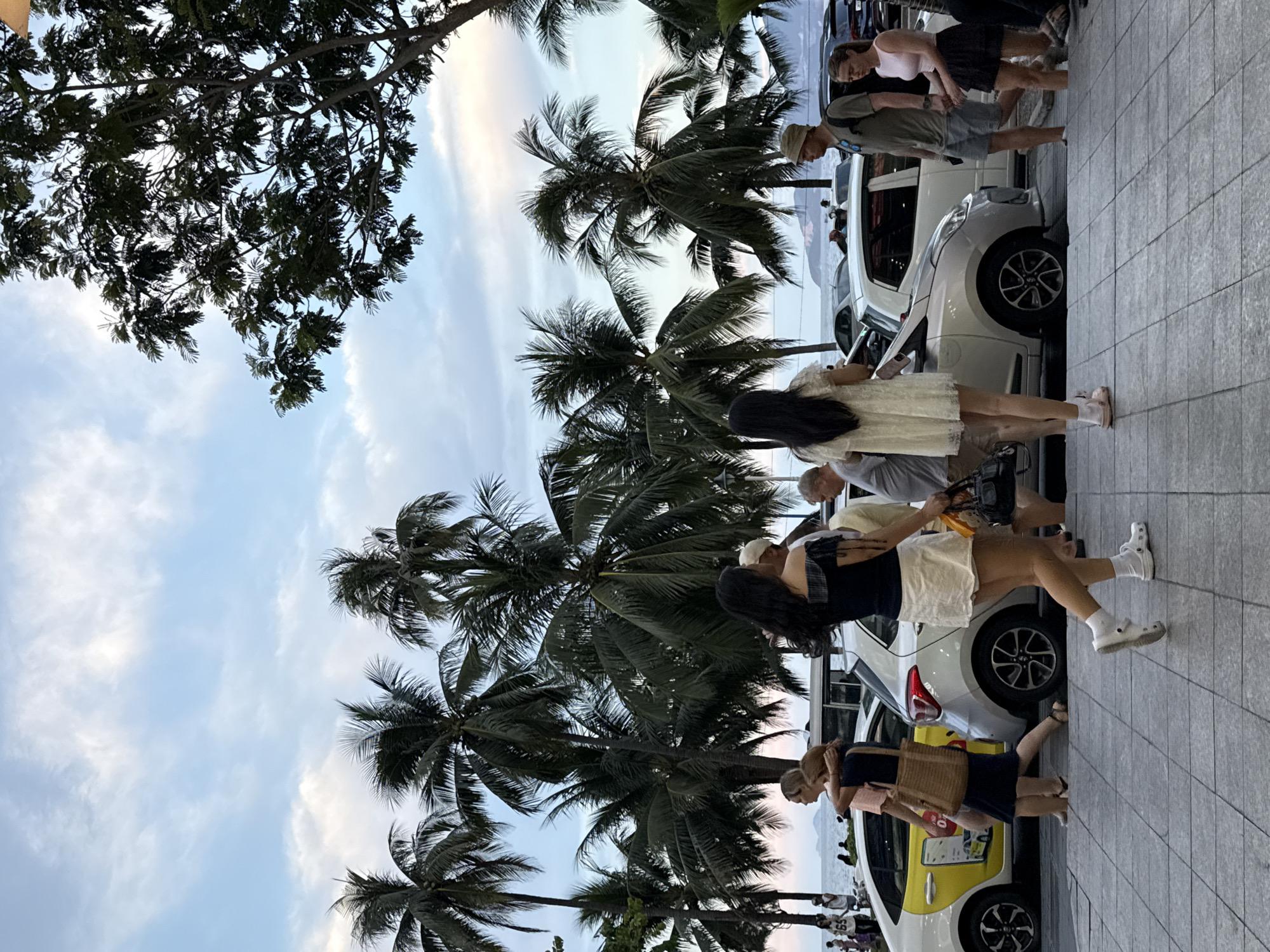The image depicts a street scene in Nha Trang Ward, Vietnam, characterized by a row of parked cars, including a yellow taxi with Vietnamese markings, and several pedestrians walking on a paved sidewalk. The background features a cluster of tall palm trees and a hazy sky with scattered clouds. The lighting suggests it is late afternoon or early evening.
In the foreground, a woman in a white dress and white Crocs walks towards the viewer's right. To her left, a man wearing a hat and gray shorts walks in the same direction. Further left, a woman in a dark off-the-shoulder top and a white skirt walks with her back to the camera. Beside her, a man and a woman are walking away from the camera, with the woman carrying a straw bag. Another woman in a dark dress and a light-colored bag is also visible.
In the middle ground, various cars are parked along the street, including a white hatchback and the yellow taxi. The taxi has a visible advertisement on its door. Beyond the cars, palm trees dominate the landscape, with the ocean and distant land visible on the horizon under a soft, pastel sky. A few people are also seen near the shoreline. The overall atmosphere is casual and hints at a coastal urban environment.