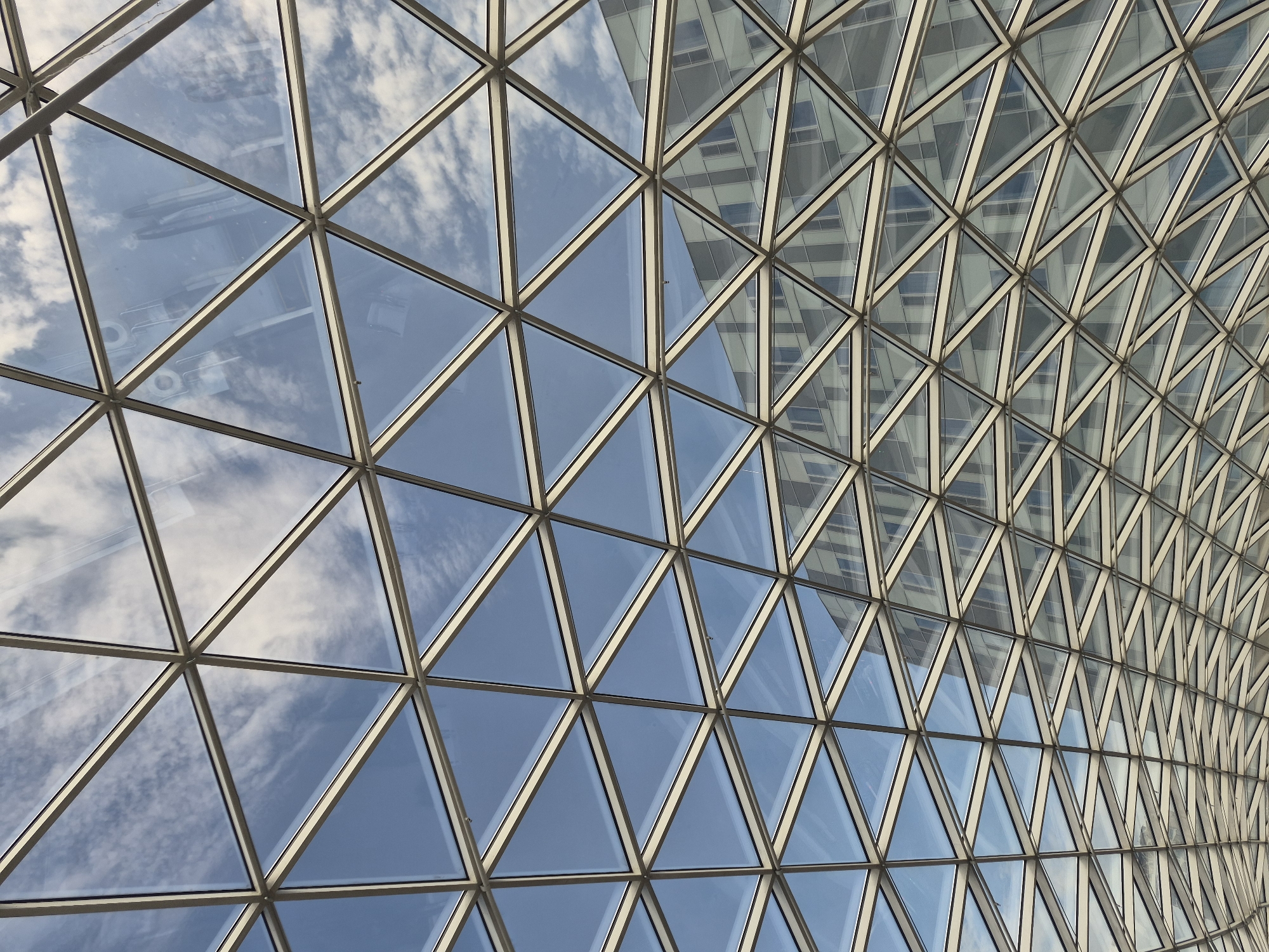 The image captures a low-angle perspective of a modern geodesic dome structure, likely a roof or atrium, with a repeating pattern of triangular glass panels framed by a light-colored metal grid. The sky is visible through the upper sections of the dome, featuring a light blue expanse with scattered white, wispy clouds. In the background, reflected in the glass panels, is a multi-story building with a facade of numerous windows and vertical architectural elements. The building appears modern, with a color palette of grays and whites. The geometric repetition of the dome's structure creates a sense of depth and visual complexity. There are no people or animals visible in the image. The scene is clean and orderly, suggesting an urban architectural setting. The overall impression is one of contemporary design and open space, with the dome serving as a transparent canopy. Based on the provided information, this architectural feature is located in the city of Khabarovsk, Russia.