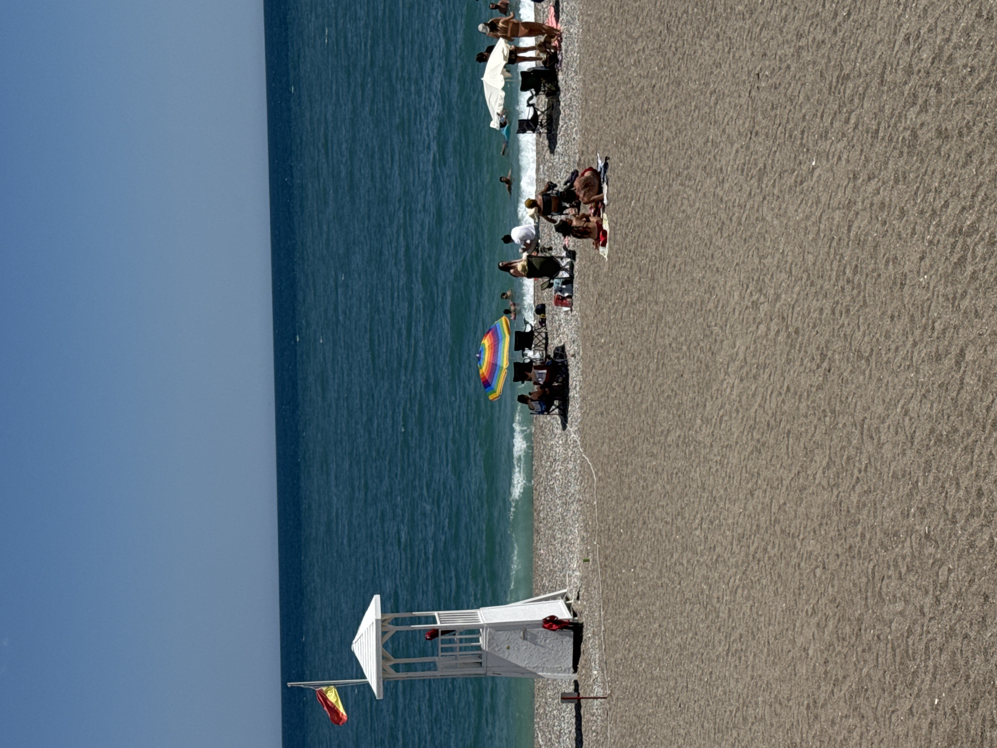 The image shows a beach scene in Konyaaltı, Turkey, taken from an elevated perspective. The beach consists of coarse, grey sand meeting the blue sea. A lifeguard station painted white stands near the edge of the beach with red rescue equipment visible inside. A Spanish flag hangs from a pole near the lifeguard station. The beach is populated with people sunbathing, swimming, and relaxing under umbrellas. There's a colorful rainbow umbrella further up the beach. The sky is clear and blue.