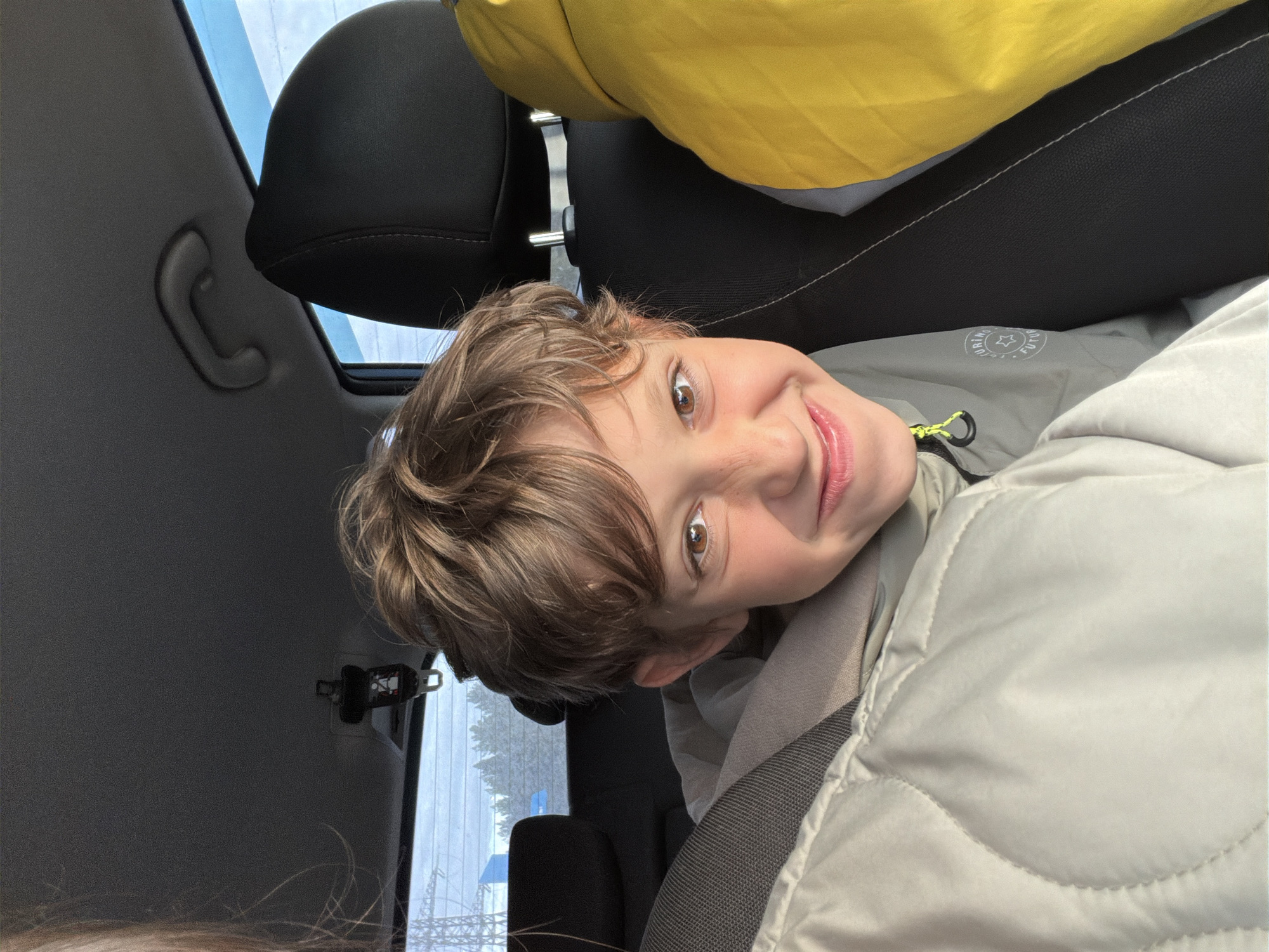 The image features a young boy with light brown, wavy hair and warm brown eyes, smiling widely at the camera from inside a car in Noviy, Russia. He is positioned in what appears to be the rear seat, looking towards the person taking the photograph. The boy is wearing a light gray or tan quilted jacket over a darker garment that has a bright yellow-green zipper pull. On his inner garment, a circular emblem is visible, featuring text that appears to read "DRIVING FUTURE" around a central star or gear symbol.
The car's interior is evident, with a black headrest behind the boy and a bright yellow object, possibly a blanket or another piece of clothing, draped over the top of the seat. Parts of the car's gray ceiling, including a handle and a seatbelt buckle near a window, are also visible. Through the window, glimpses of an outdoor scene can be seen, suggesting daytime conditions and possibly bare trees or distant power lines, hinting at a natural or semi-urban environment typical of Noviy, Russia. The boy's cheerful expression conveys happiness, and his attire suggests cooler weather, possibly during the winter months. The image captures a moment of joy during a car journey.