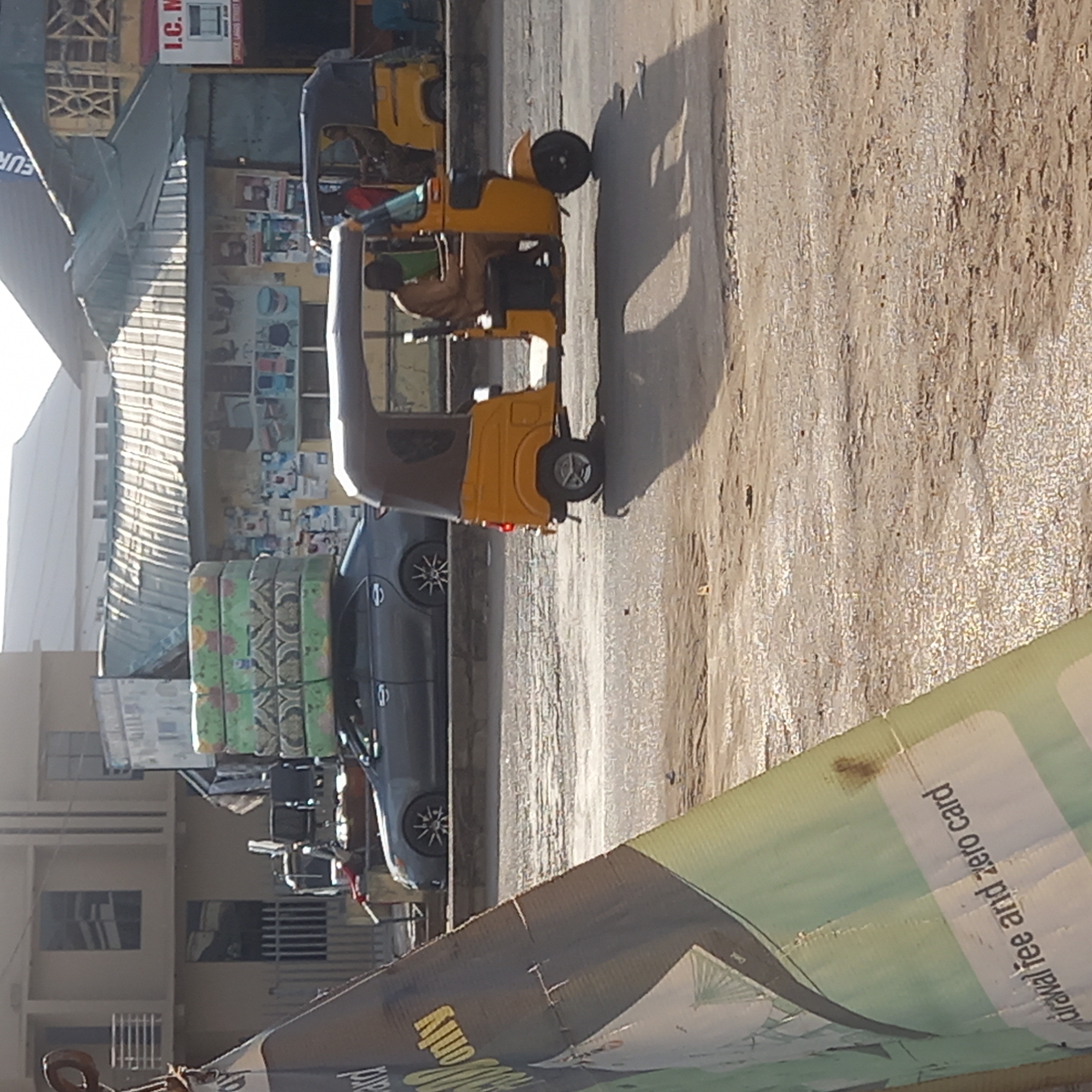 The image shows a street scene in Sardauna, Nigeria. Three yellow auto-rickshaws, known locally as "Keke Napep," are parked on the side of a dusty street. A man can be seen inside the first vehicle. Beside the rickshaws sits a grey sedan, parked in front of a building with mattresses stacked on top of each other. In the foreground, there's a damaged advertising banner. The environment seems urban with buildings visible on the side of the image.