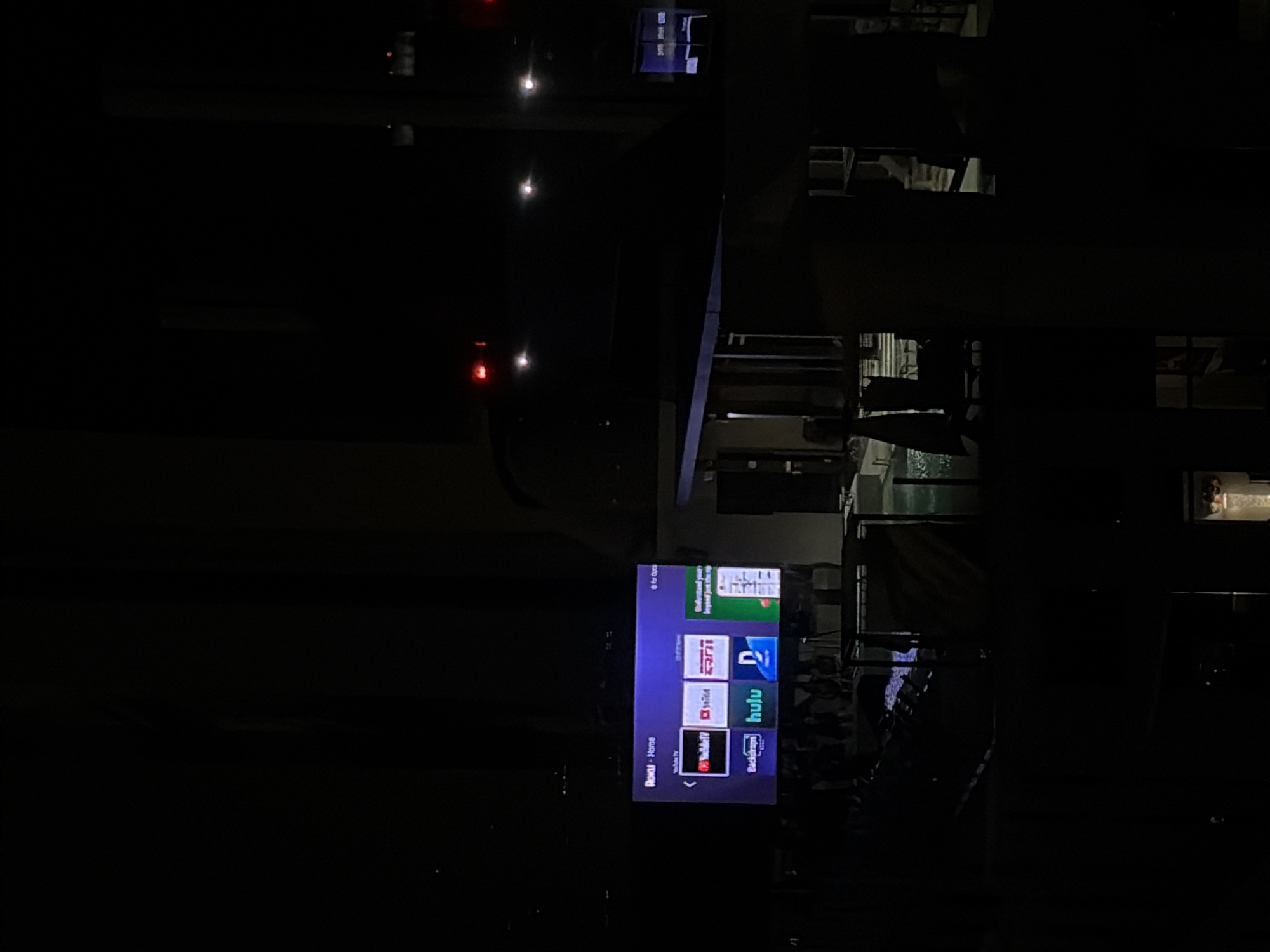 The image shows a television displaying the Roku home screen, with icons for YouTube TV, Hulu, ESPN, and other streaming services. The television appears to be located outside, likely on a balcony or patio. In the background, there is a view of city buildings and streetlights. Based on the available information, it's not possible to pinpoint the exact location within Austin.
