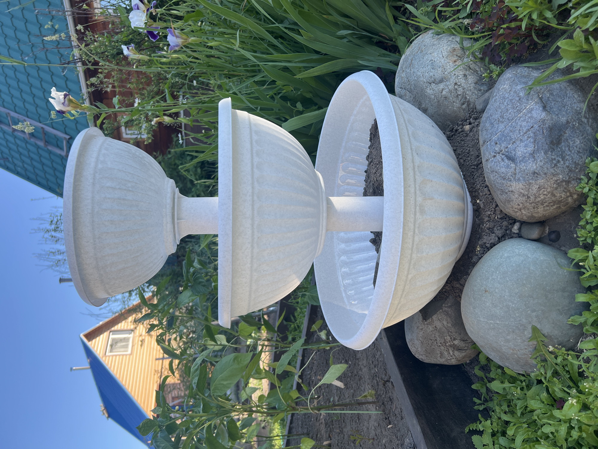 A tiered, off-white plastic planter sits in a garden on a sunny day. The planter has three bowls of decreasing size, stacked on a central pole, with the bottom bowl filled with soil. Large, smooth grey rocks surround the base of the planter, nestled amongst green vegetation and some dark soil. In the background, a rustic wooden house with a dark green metal roof and a wooden house with an orange exterior and a bright blue metal roof are visible. Green plants, including what appear to be pepper plants and iris flowers, are growing around the planter and between the houses. The sky is a clear, bright blue. The scene is located in Mochische, Russia.