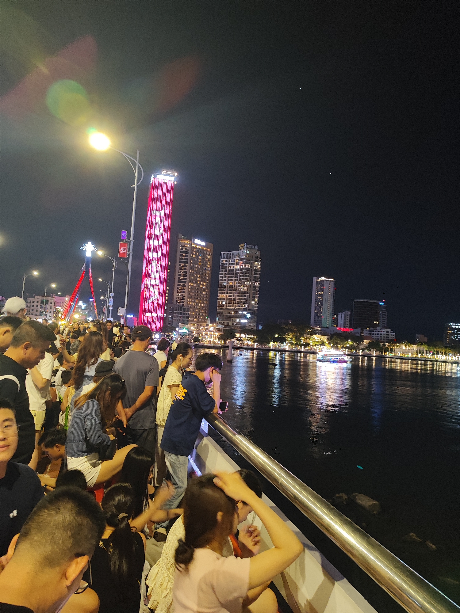 At night, a crowd of people is gathered alongside a bridge in Đà Nẵng, Vietnam. The bridge has a silver railing. Behind the crowd are several illuminated buildings, including one tall building with red lights, the Hilton Đà Nẵng. In the water, a boat is visible and illuminated. The environment includes a night sky.