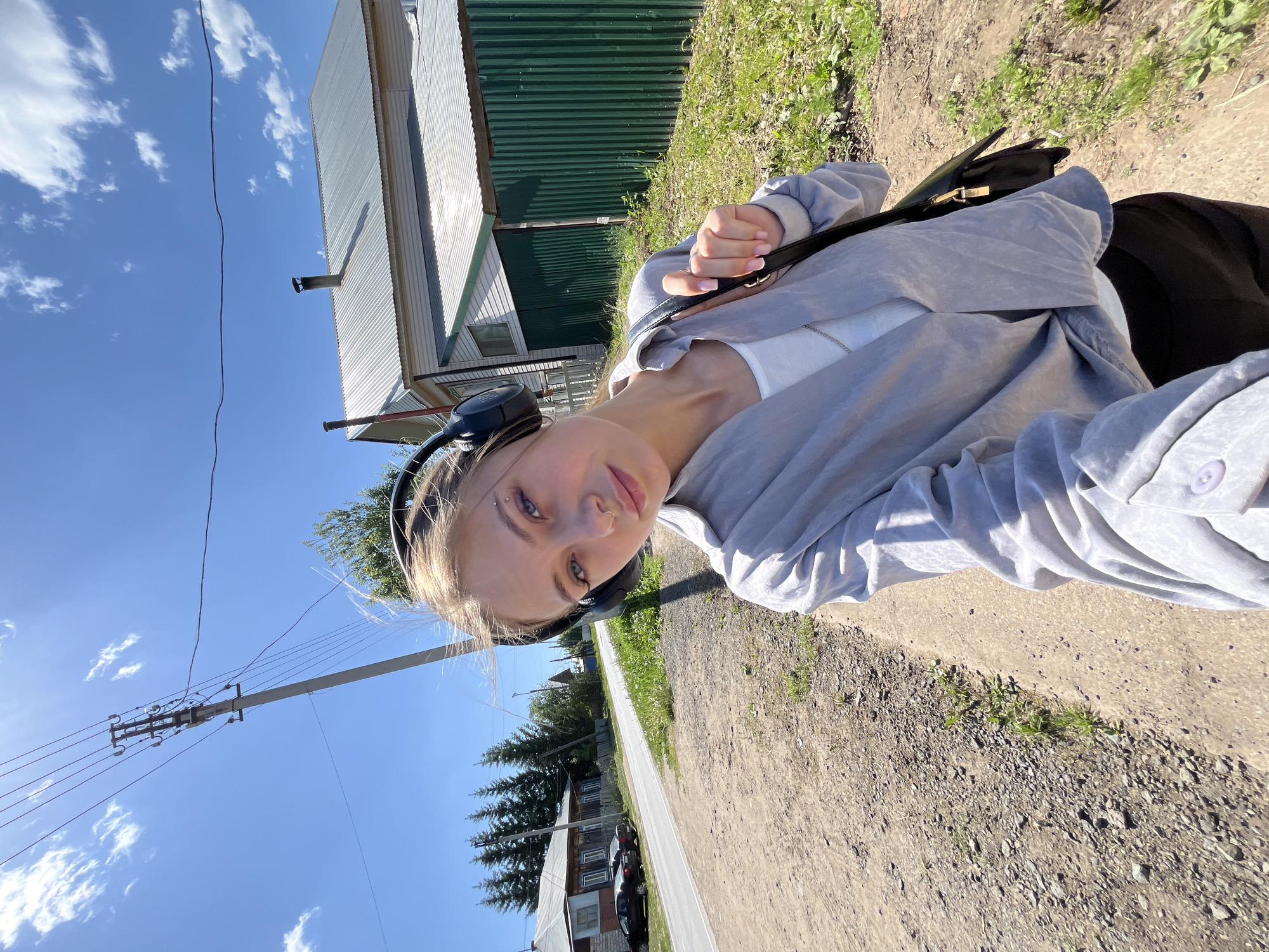 A young woman wearing headphones, a grey jacket and a white top is outdoors, possibly on a sidewalk or paved area next to a road. She has blonde hair and light skin. She is carrying a black purse or bag over her shoulder. In the background are houses, trees, and a utility pole. The sky is blue with scattered clouds. The location is Фоки, Russia.