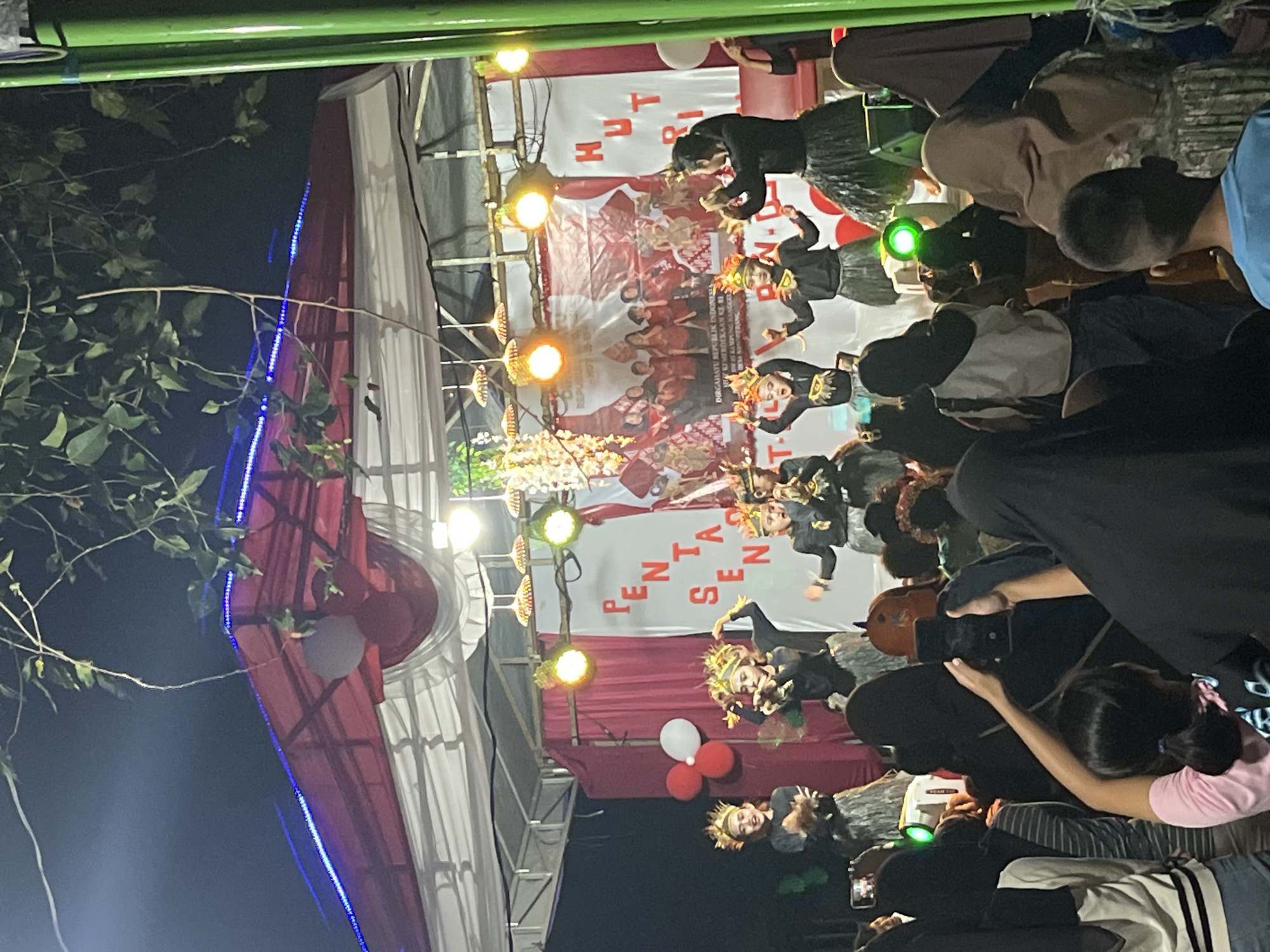This is a full-shot, vertical, eye-level outdoor photograph taken at night in Tajurhalang, Indonesia. The image captures a cultural performance on a stage. In the center of the frame, several young girls, dressed in traditional attire with elaborate headpieces, are performing a dance. They wear black tops and skirts, with some accessorized with feathery elements. Behind them, a banner with red and white fabric and text that appears to read "PENTAS SENI HUT RI" is displayed. Several lights illuminate the stage, including hanging decorative lights and spotlights. The stage itself is covered by a red and white tent structure, adorned with blue LED lights. In the foreground and to the sides, a crowd of spectators watches the performance. Many in the audience are holding up their phones, presumably to record the event. The overall atmosphere suggests a community celebration or festival.