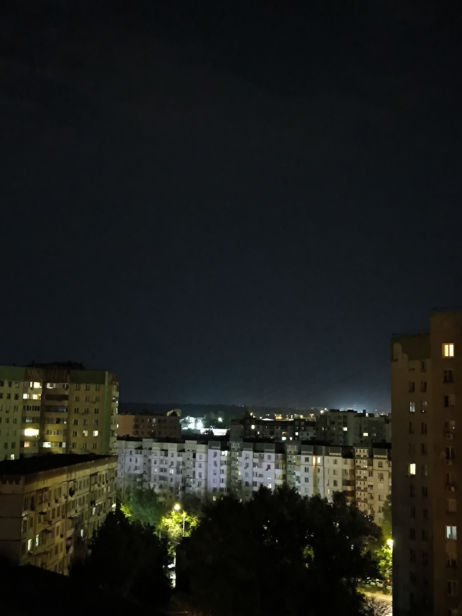 The image depicts an urban landscape at night, likely in Chișinău, Moldova. The scene is dominated by a dark night sky, with a faint glow on the horizon indicating distant city lights. In the foreground and midground, several multi-story apartment buildings are visible. These buildings are characteristic of Soviet-era architecture, with rows of identical windows, some of which are illuminated, suggesting occupancy.
On the left side of the image, a building with a greenish-yellow facade and balconies is prominent. To its right, a cluster of pale, multi-story buildings forms the central part of the cityscape. These buildings also have numerous illuminated windows. In the far right, a taller, brownish building with a simpler facade stands out. The lower parts of the image are filled with dark silhouettes of trees, interspersed with the glow of streetlights. The overall impression is one of a typical residential area at night, with the characteristic lighting of apartment blocks.