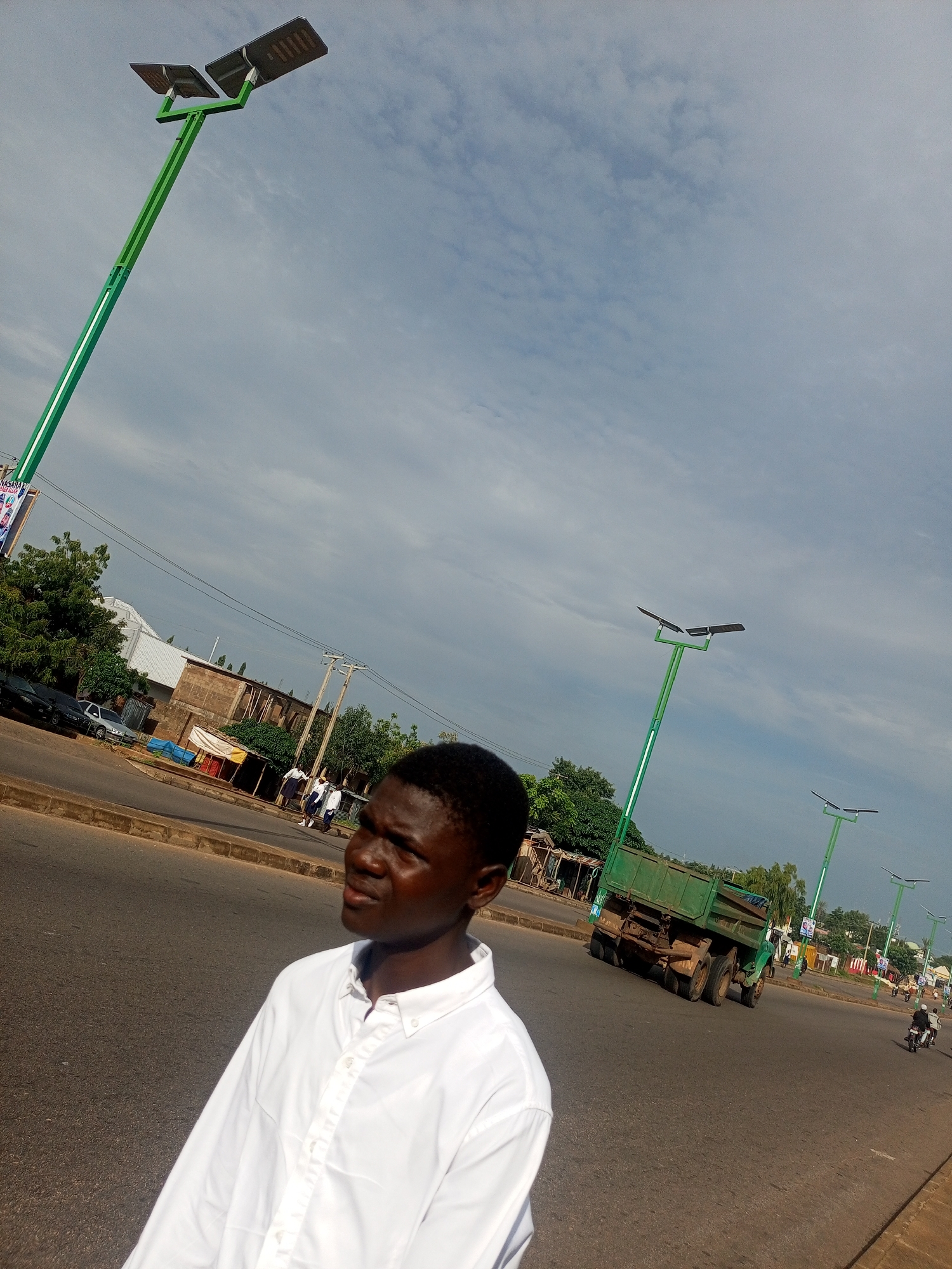 The image depicts a street scene in Tempure, Nigeria. In the foreground, a young man wearing a white button-down shirt is looking to his right. The background features a road with several vehicles, including a green truck in motion, and a line of tall green lampposts, some of which are equipped with solar panels. Buildings and trees are visible along the side of the road. The sky is cloudy.