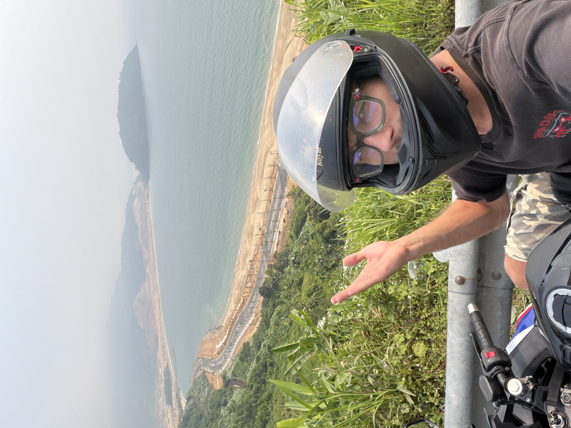 A man wearing a black motorcycle helmet and glasses is taking a selfie overlooking a coastal scene. In the background, there's a body of water with a land formation in the distance, possibly an island. The shoreline features a man-made structure or road, and vegetation covers the terrain leading down to the coast. The image is slightly angled, with the person and the front of a motorcycle appearing in the foreground on the right side. The location is likely near the city of Da Nang, Vietnam based on the landscape and coastal features visible.