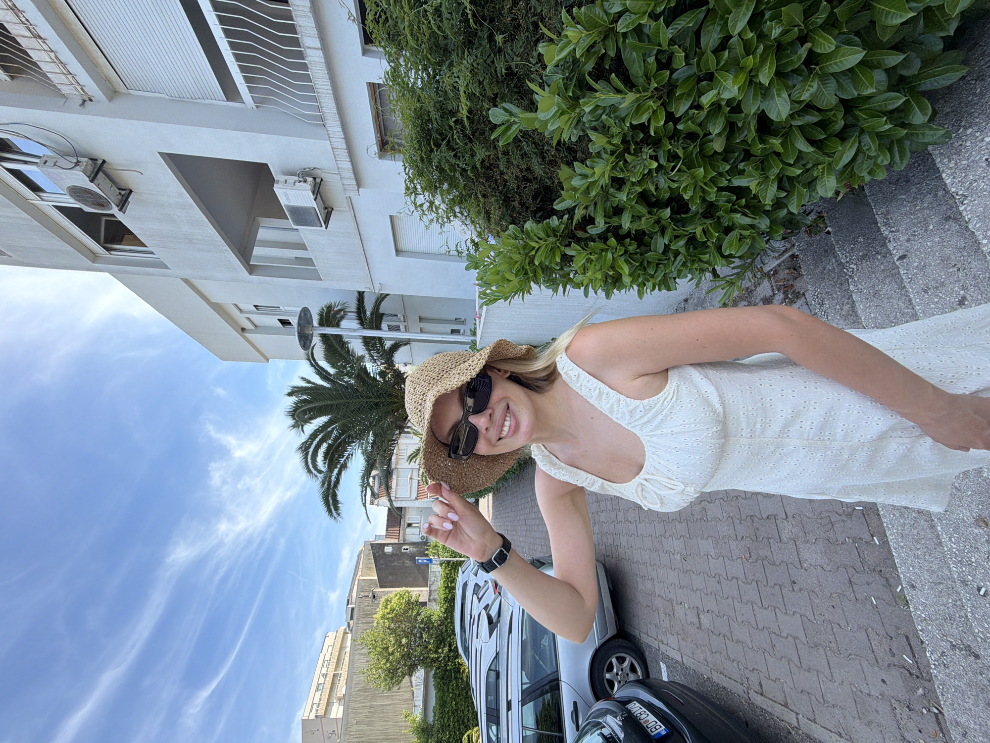 A blonde woman wearing a white eyelet dress and a straw hat smiles while holding the brim of her hat. She is standing on a set of concrete stairs in Budva, Montenegro, with a white building and lush green bushes behind her. In the background, a parking lot with several cars and palm trees are visible under a blue sky with wispy clouds.