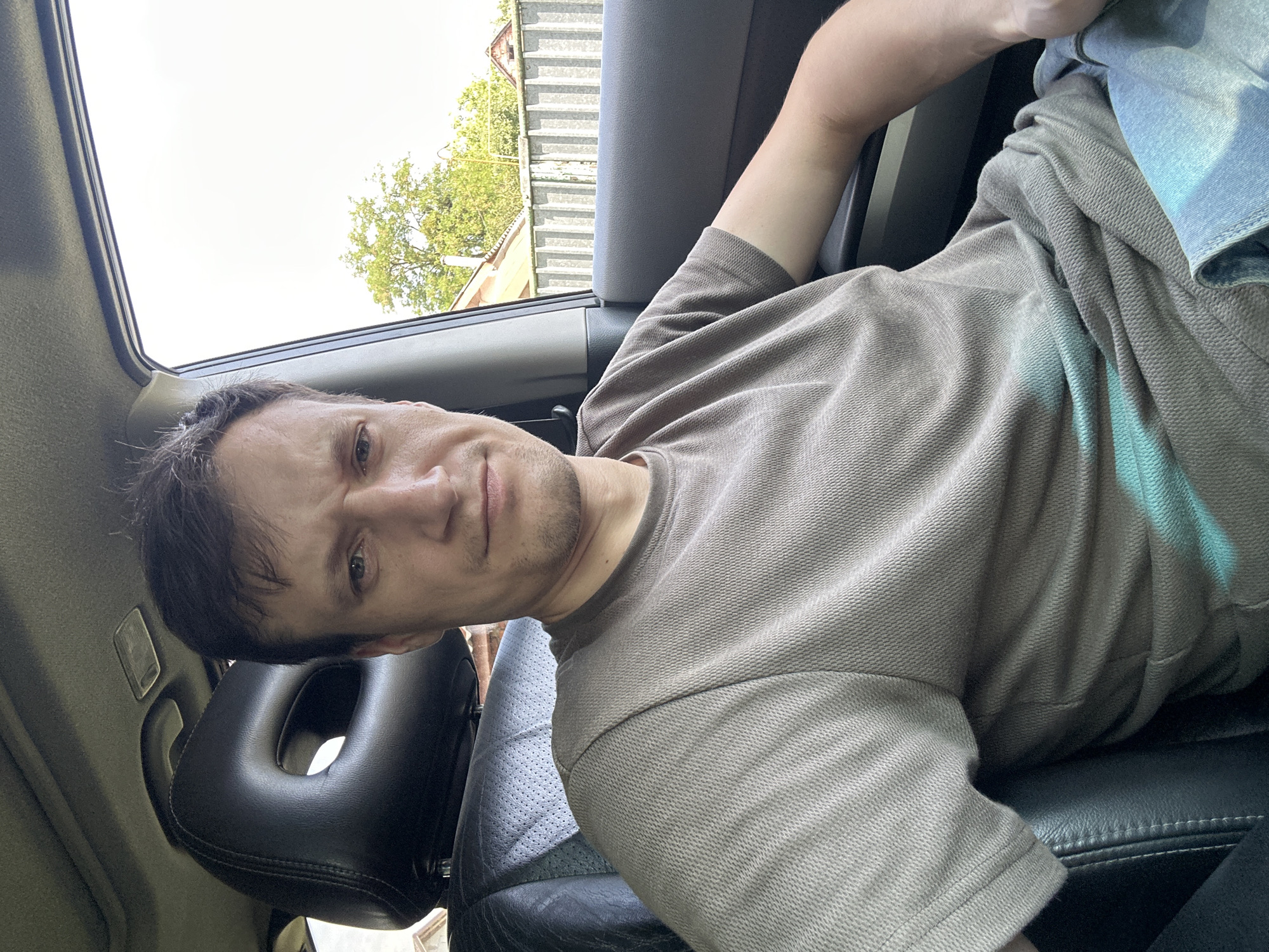 A man is sitting in the driver's seat of a car, looking directly at the camera. The car appears to be parked, as indicated by the stationary background. The man is wearing a gray t-shirt and light blue jeans. The interior of the car is visible, including the headrest and part of the dashboard. Through the window, houses and trees can be seen, suggesting an outdoor location, possibly in a residential area. The overall lighting suggests it is daytime.