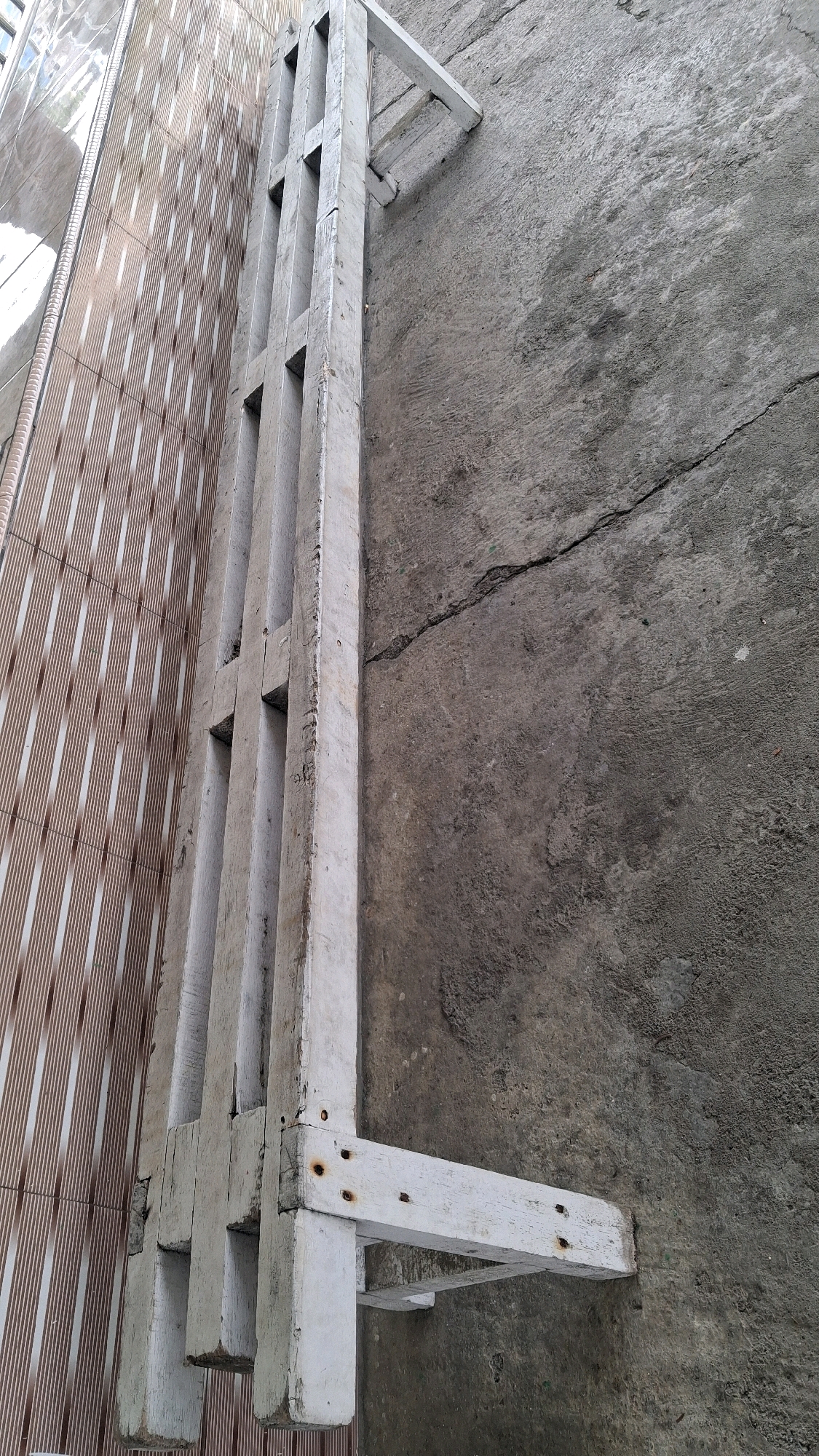 The image shows a section of an exterior wall with a wooden structure attached to it. The wall on the left is covered in vertical striped tiles, appearing to be light brown and white. To the right of the tiled wall is a rough, gray concrete surface with a prominent crack running horizontally across it. Attached to the concrete wall is a white, wooden structure that appears to be a ladder or a series of shelves. This wooden structure is made of planks and has a somewhat weathered appearance with visible screws and nails. The image is taken from a low angle, looking upwards. The overall scene suggests an outdoor environment, possibly a side alley or a backyard in a location such as Giriwarno, Indonesia, given the common architectural elements and materials. There are no people or animals present in the image.