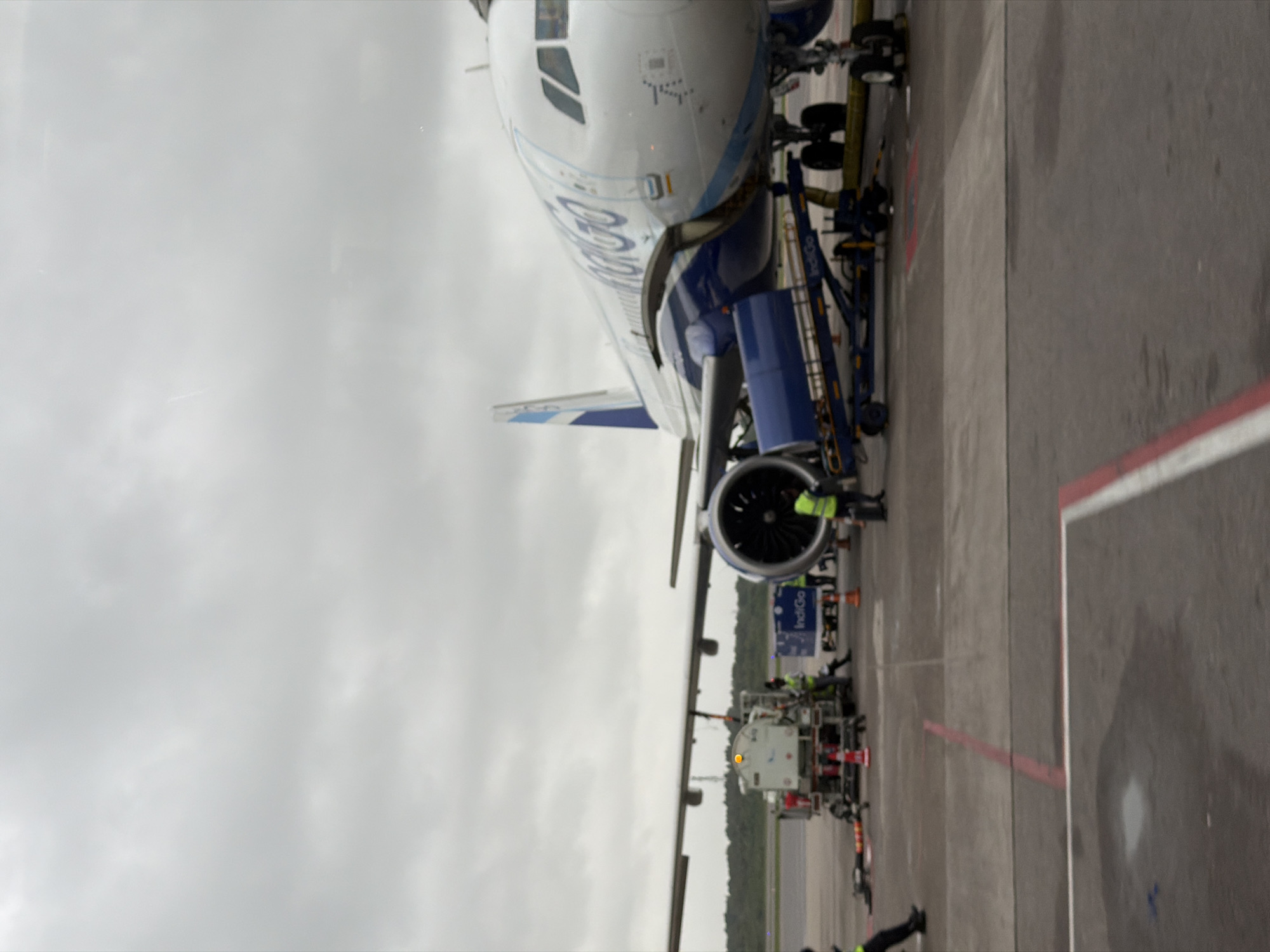 The image depicts an Indigo Airlines aircraft parked at an airport tarmac on an overcast day. In the foreground, the tarmac is marked with red and white lines. Several ground crew members in high-visibility vests are attending to the aircraft. To the left, a fuel truck is connected to the plane, with an orange warning light visible. A baggage conveyor belt, also bearing the Indigo logo, is positioned near the aircraft's cargo door. The aircraft itself is predominantly white with blue accents and the distinctive Indigo branding. The background shows a line of trees and a cloudy sky, characteristic of the general atmosphere at Rajiv Gandhi International Airport in Hyderabad, India, during a potentially humid and temperate day.