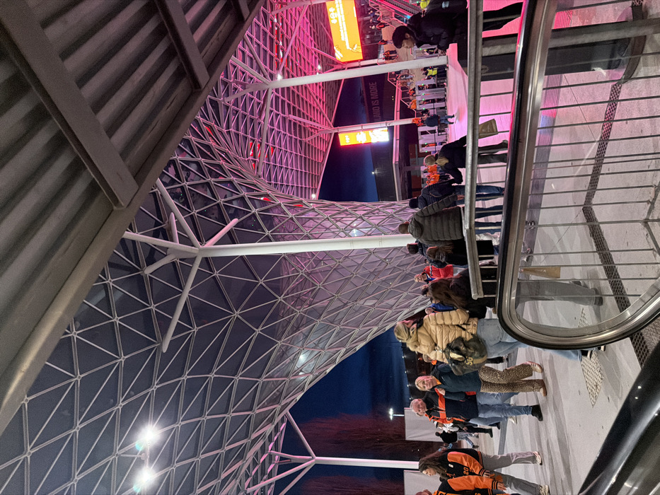 People stand in line next to a moving walkway. The image is tilted, showcasing an intricate, triangular-patterned glass and steel architectural structure overhead. Part of a corrugated metal roof is visible to the side. The structure is illuminated with pink and white lights, and nearby buildings have signs. The sky is dusky blue.