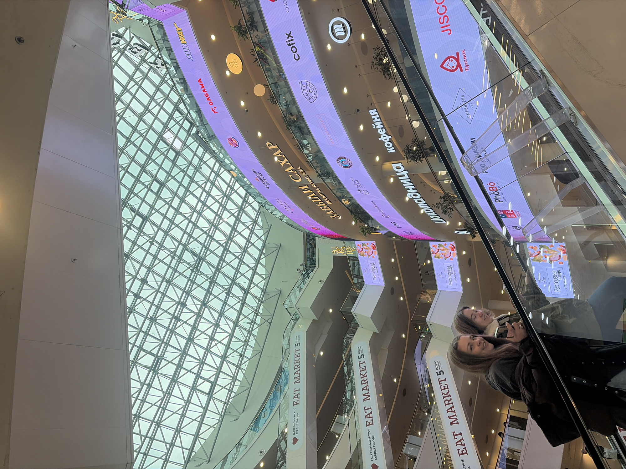 This image captures the interior of a spacious, modern shopping mall, likely located in Saint Petersburg, Russia. The scene is dominated by a vast, intricate glass and metal grid ceiling structure, resembling an atrium, which allows ample natural light to illuminate the space, suggesting it is daytime.
In the foreground, two women are riding an escalator. One woman, with long brown hair, smiles towards the camera while holding a smartphone, her reflection clearly visible in the escalator's glass balustrade. Beside her, a second woman with shorter, lighter hair is also engrossed in her phone. Both appear relaxed.
The mall features multiple curved levels, each adorned with illuminated digital signage panels showcasing various food and beverage brands. Below these panels, large vertical banners are displayed, repeatedly featuring "EAT MARKET 5" and the Russian text "ГАСТРОНОМИЧЕСКОЕ СЕРДЦЕ ГОРОДА" (Gastronomic Heart of the City), alongside "Рестораны и кафе" (Restaurants and cafes), indicating a significant dining area.
Numerous brand names are visible in both English and Russian, including international chains like SUBWAY, BURGER KING, and COFIX. Russian brands and phrases include "Teremok," "Shokoladnitsa" (Шоколадница), "кофейня" (coffee shop), "ЗАБЫЛИ САХАР" (Forgot Sugar), "MACARONIKA," "PhoBo" (with "ВЬЕТНАМСКАЯ КУХНЯ" - Vietnamese Cuisine), GAGAWA, PIMS, SURE COFFEE, JISCO, VRAVAWE, ФРАНКЛИНс (Franklins), and СЫРОВАРНЯ (Cheese Factory/Dairy). The overall impression is that of a bustling, architecturally impressive commercial hub offering a wide array of dining choices.