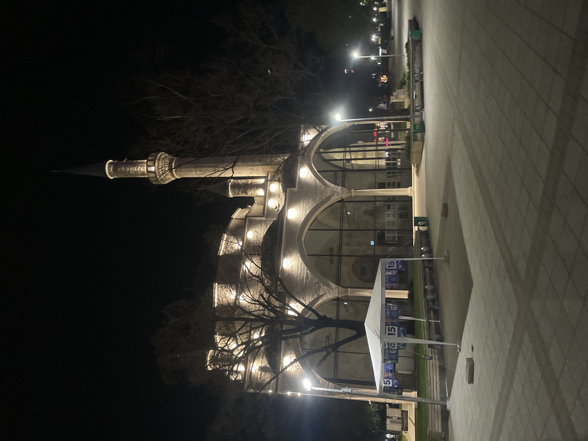 The image shows the Ibrahim Pasha Mosque in Razgrad, Bulgaria, at night. The mosque is illuminated, highlighting its architectural features, including the minaret and arches. A bare tree stands in front of the mosque. In the foreground, a white tent-like structure is visible with banners or signs attached to it. A paved area is in the foreground and side of the image, with streetlights and other urban elements visible in the background. The scene appears to be a public space or square.