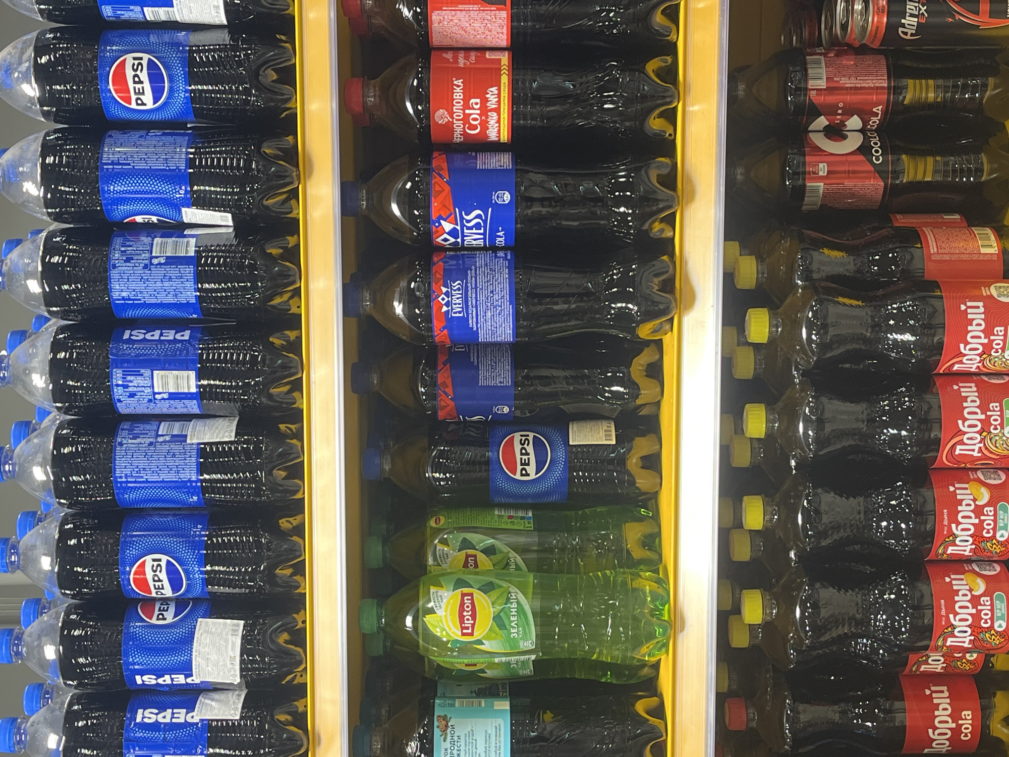 The image displays a refrigerated shelf in a supermarket or store, filled with various bottled and canned beverages. The products are arranged in vertical rows, mostly large plastic bottles of cola and tea.
On the left side of the frame, there is a prominent section dedicated to Pepsi, featuring numerous large bottles with blue labels. The word "PEPSI" is clearly visible on the labels.
The middle section of the display showcases a variety of brands. At the top, there are bottles of "ЧЕРНОГОЛОВКА Cola" (Chernogolovka Cola) with red labels, some showing the text "IMITRIENKO VANYA". Below these are bottles of "EVERVESS COLA" with distinctive blue and red labels, marked as "ВИТОК БЕЗАЛКО" (likely part of "Безалкогольный," meaning non-alcoholic). Further down, more Pepsi bottles are visible, followed by several bottles of "Lipton ЗЕЛЕНЫЙ ЧАЙ" (Lipton Green Tea) with green labels. Another label mentions "TOK ПРОДНОЙ ЖЕСТИ".
The right side of the display features additional beverage options. At the top, a can of "Adrenaline EXT" (Adrenaline Rush energy drink) is partially visible. Below it are bottles of "COOL COLA ZERO" with red and black labels, featuring a large "C". The lower part of this section is dominated by multiple bottles of "Добрый cola" (Dobry cola) with red labels, some specifically indicating "Вкус Дыня" (Melon flavor) and "HIP HOP".
The products are neatly stacked, indicating an organized retail environment. All visible text is in Cyrillic, consistent with the location context of Saint Petersburg, Russia.