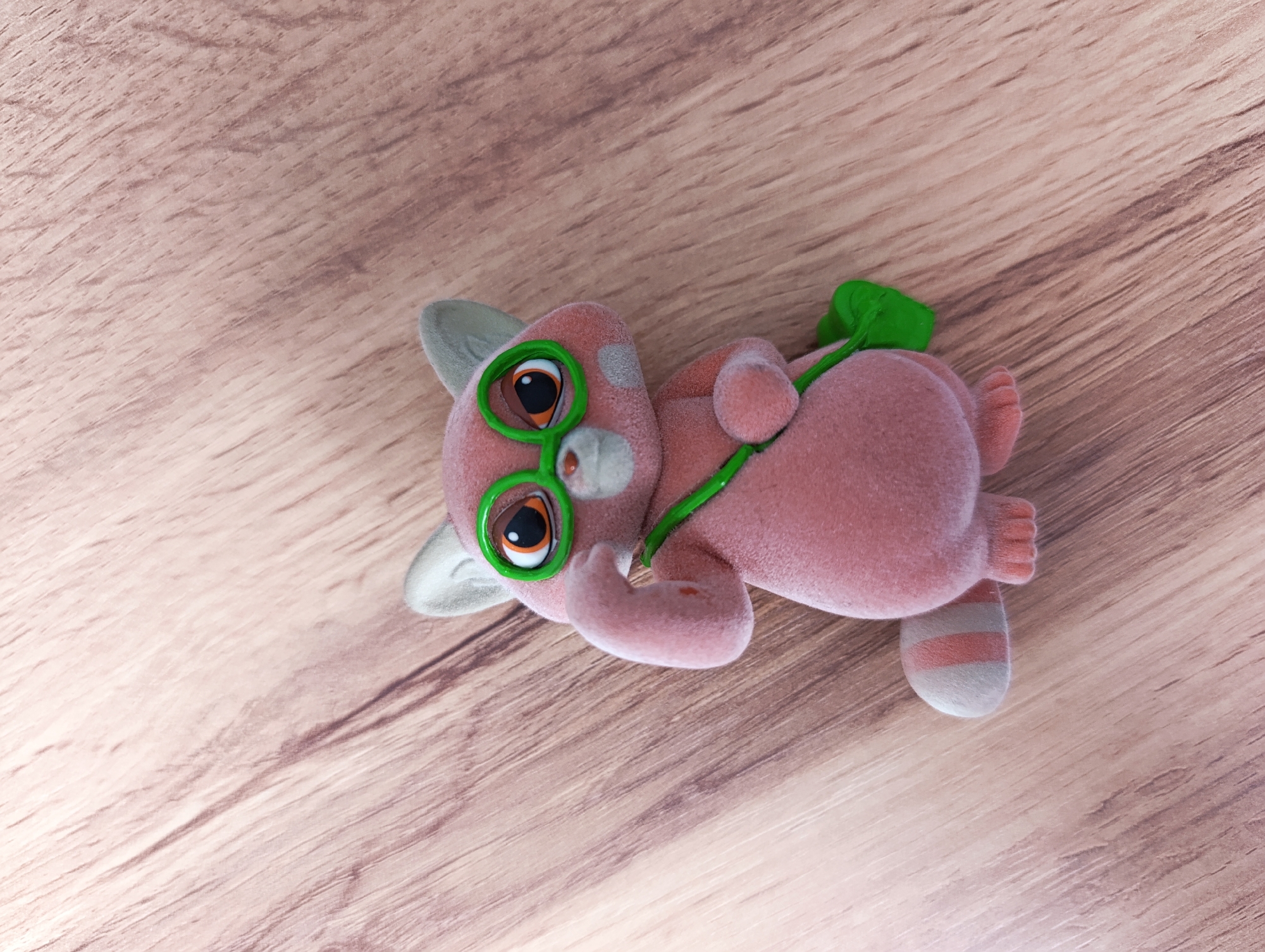 A small, pink, flocked animal figure is depicted lying on its left side on a light-colored wooden surface. The figure, possibly a cat or a similar creature, features large, round eyes with bright green rims and dark pupils, along with small, white pointy ears and a tiny white nose. A thin, bright green strap or ribbon, resembling a vine or leaf stalk, is draped across its back and wraps around its body. Its body is primarily pink, with a visible striped pattern of white and pink on one of its back legs. The wooden surface displays a subtle grain pattern, suggesting an indoor setting like a floor or table. The lighting is bright and even, indicating daytime. No human subjects or text are present. The image captures a still-life of the whimsical toy.