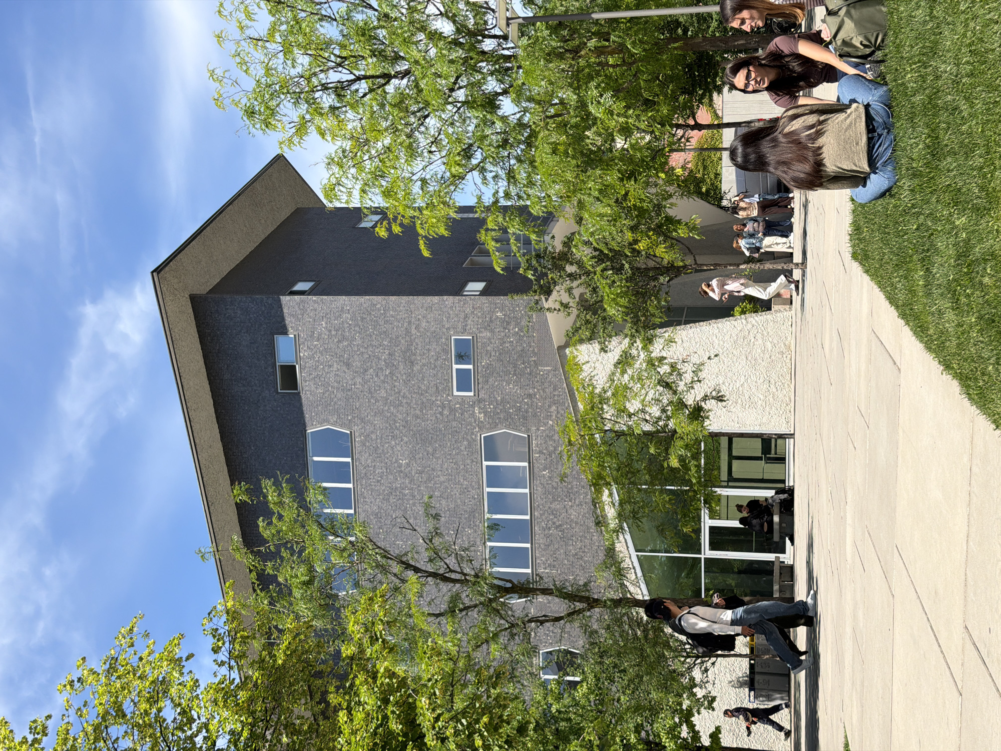 The image depicts a building with a dark gray facade and a sloping roof in an outdoor setting. The building has multiple windows, including some arched windows. Trees with green foliage are present in the foreground, partially obscuring the view of the building. People are visible on a walkway adjacent to the building, some are sitting on the grass while some are walking or standing near a glass entrance. A paved area is also visible. The sky is blue with some clouds. The image is rotated sideways. The location appears to be an academic setting, possibly in Milan, Italy.