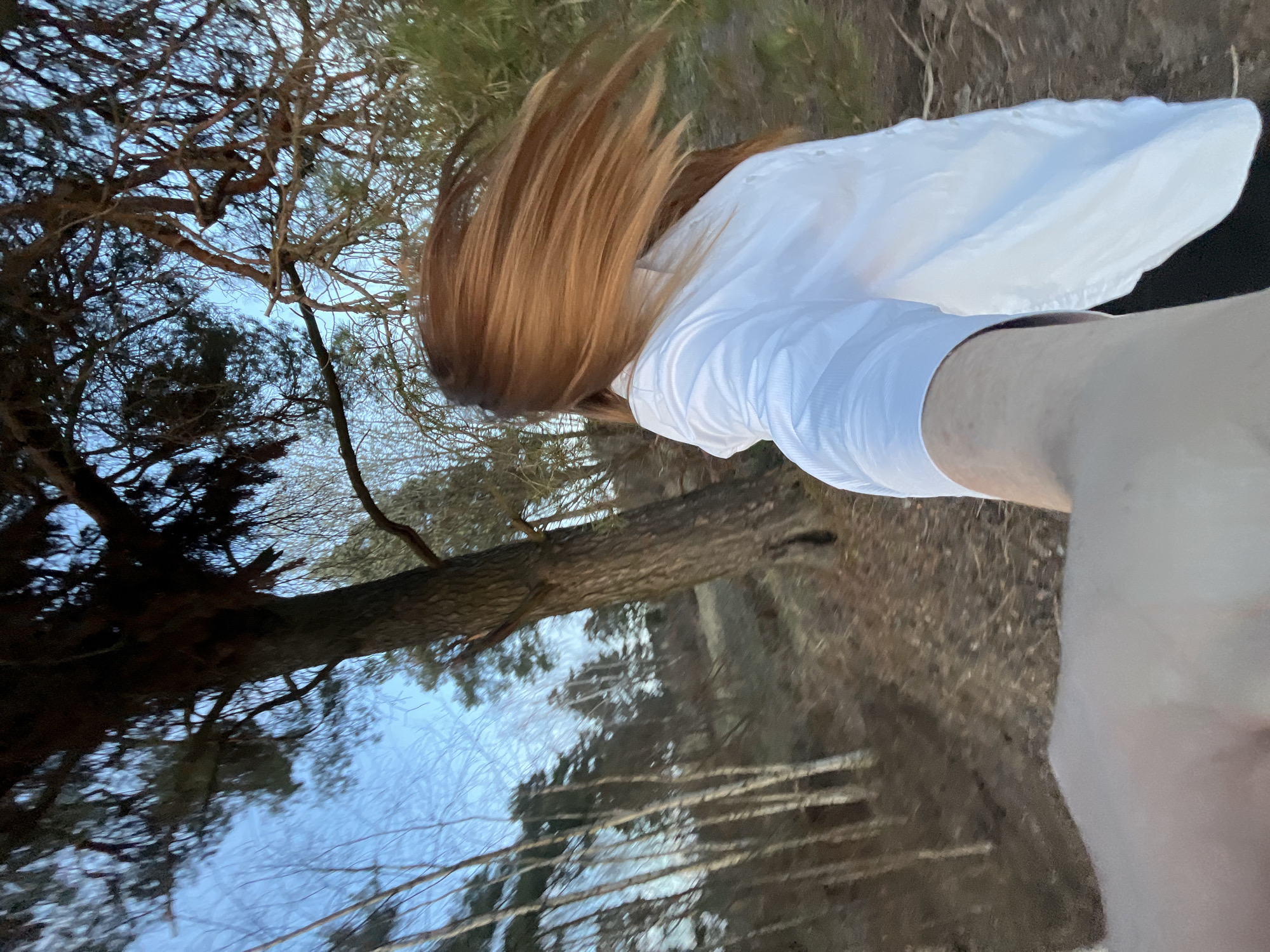 A woman with long, reddish-brown hair wearing a white top is standing in a forest in Razdolye, Russia. The ground is covered in brown grass and pine needles. Trees are visible in the background, silhouetted against a blue sky.