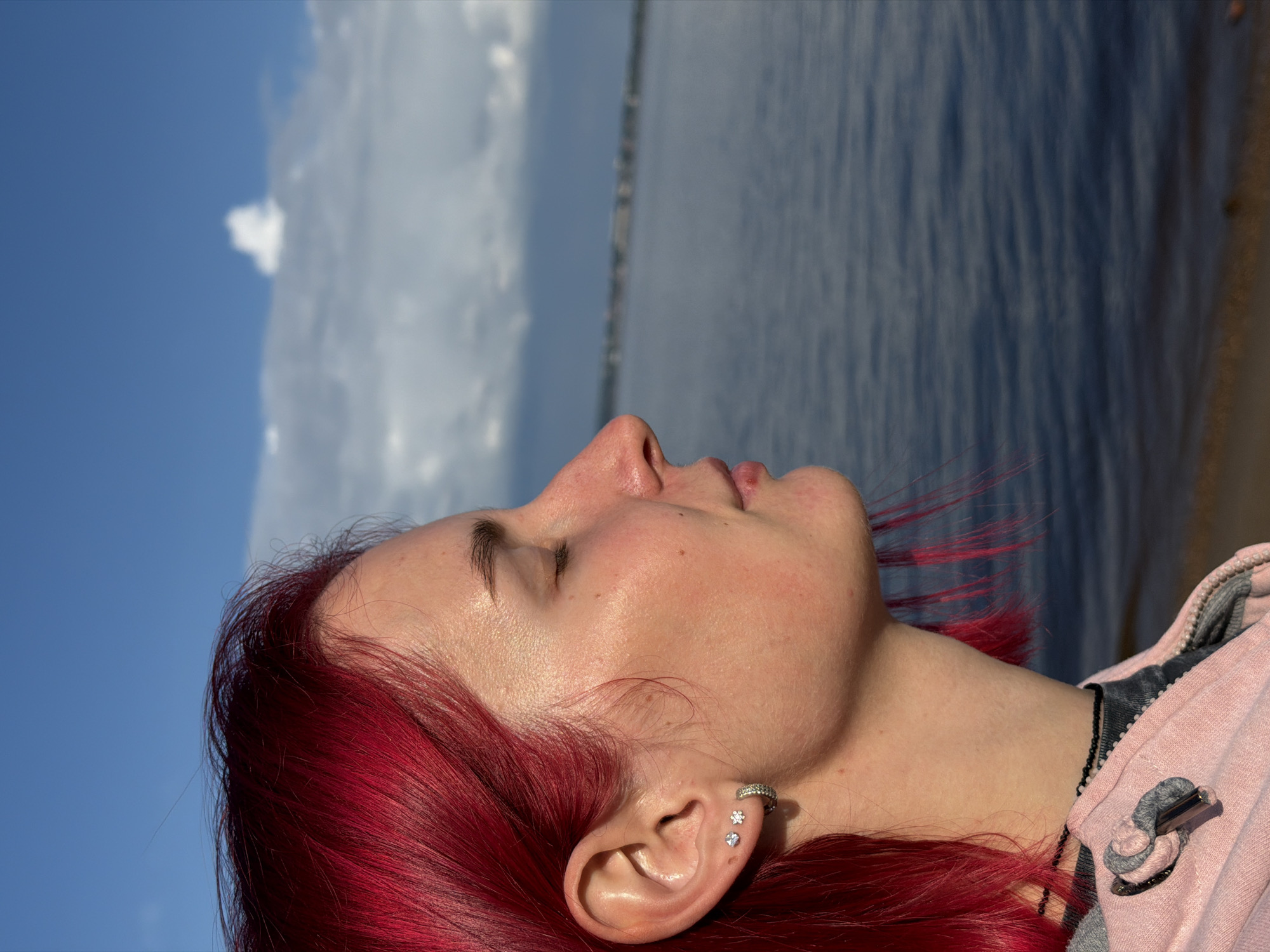 This close-up, sideways portrait features a young adult with striking bright red hair, positioned against a clear blue sky and a calm body of water. The subject's head is tilted upwards, eyes closed, and face in profile, suggesting a moment of relaxation or contemplation while basking in the sunlight. Sunlight highlights her skin, particularly on her forehead and cheek. She is wearing a light pink hoodie or jacket, with a black zipper and some grey detailing. Her visible ear is adorned with several piercings: an ear cuff with small crystals, a flower-shaped stud, and a round crystal stud. A thin black beaded necklace or chain is visible around her neck, disappearing into her collar.
The background shows a division between the expansive blue sky on the left, dotted with soft white clouds, and the shimmering surface of a body of water, possibly a lake or sea, on the right. A faint horizon line marks the separation. The image conveys a peaceful, outdoor scene during daylight hours in what appears to be a natural setting, consistent with the location context of Noviy, Russia. There is no visible text in the image.
