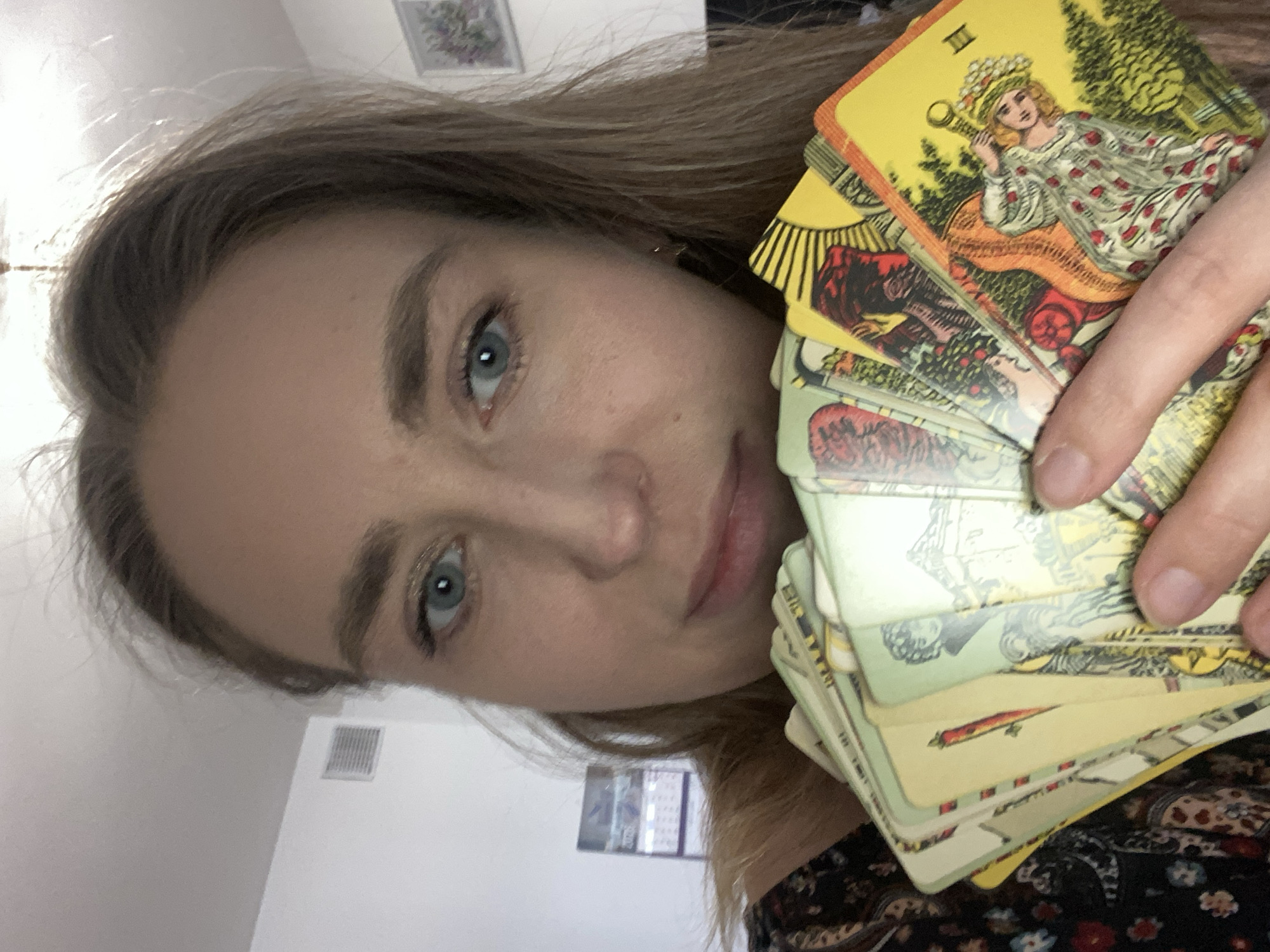 A woman with light brown hair and blue eyes holds a stack of tarot cards. The top card depicts a woman in a crown, seated on a throne. Other cards in the stack feature imagery of animals and figures. The background is a white wall with a framed picture. The woman is wearing a dark-colored floral print top. The location is not apparent from this image.