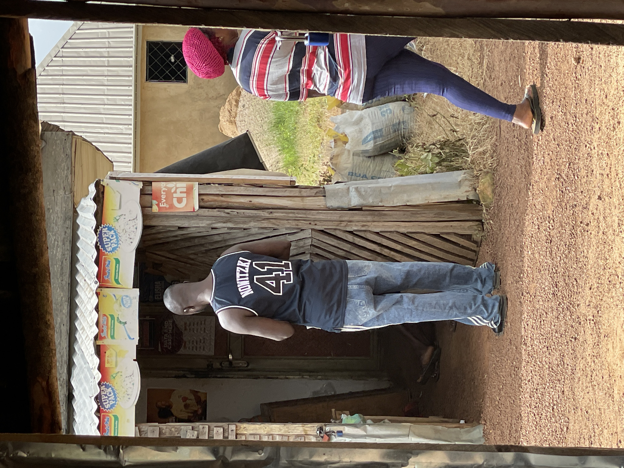In Durumi, Nigeria, a scene unfolds outside a small, open-air shop. The shop, constructed with a corrugated metal roof, displays various colorful packets of "Indomie Instant Noodles" and "Super Pack" products above its entrance. A poster featuring an image of a person in a yellow shirt is visible on the left side of the entrance.
A man, seen from the back, stands facing the shop. He is wearing a dark blue sleeveless jersey with "NOWITZKI 41" printed in white lettering and light blue denim jeans. He appears to be interacting with someone or something inside the shop.
To the right of the entrance, a woman walks away from the shop. She is wearing a bright pink head covering, a horizontally striped shirt in red, gray, and black, and dark blue leggings. Her posture suggests she is moving out of the frame.
Behind the shop, a building with a light brown stucco exterior and a silver corrugated metal roof is visible, featuring a small window with a diamond-shaped grill. Piles of sand and bags of what appear to be cement are located in the background. The ground is covered with reddish-brown gravel. The overall environment suggests a rural or semi-urban setting.