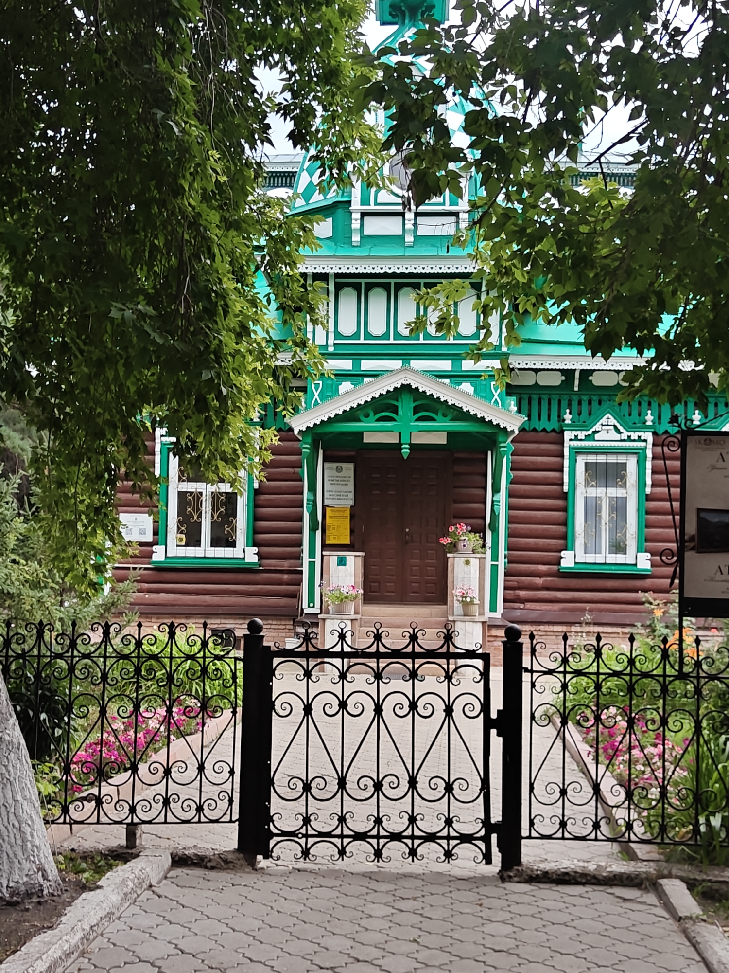 The image depicts the exterior of a historic wooden house, likely a museum, in the city of Petropavl, Kazakhstan. The house is painted in vibrant green and brown, with intricate white wooden trim adorning the roofline and windows. A dark brown wooden door is visible at the entrance, flanked by flower pots. A black wrought-iron fence with decorative scrollwork stands in front of the property, partially obscuring a glimpse of the garden beyond, which features pink and white flowers. Paved pathways lead to the entrance. The setting suggests a sunny day, with lush green foliage from nearby trees framing the top of the scene.