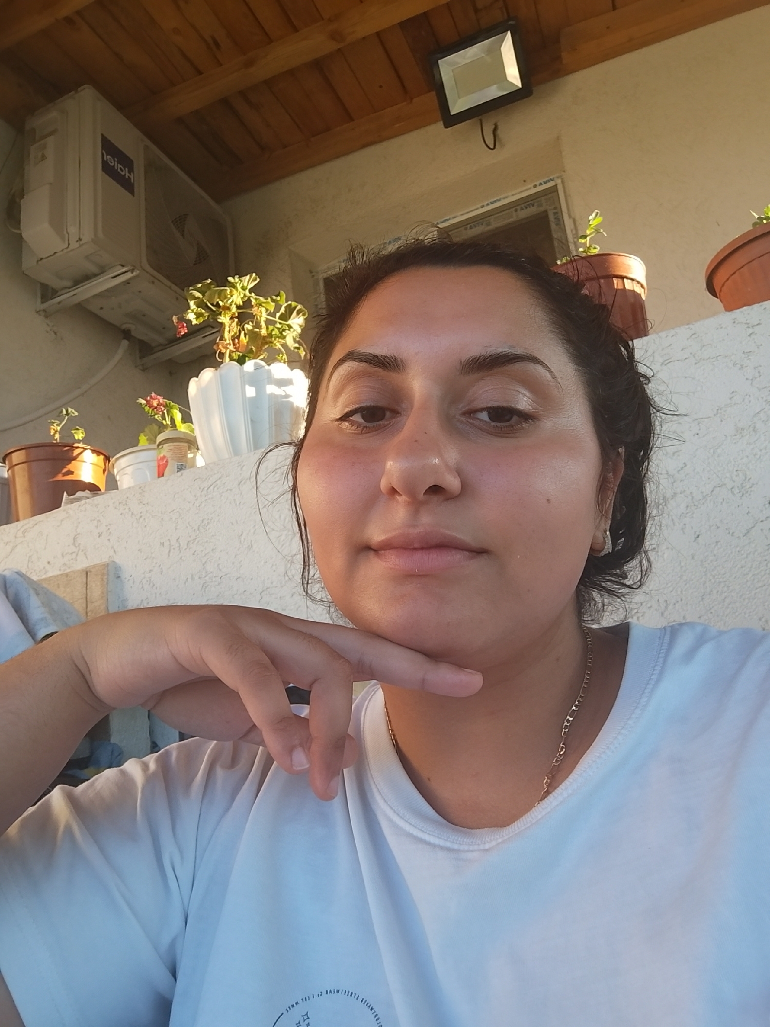 A young woman with dark hair is pictured from the chest up, outdoors, possibly on a patio or balcony. She is resting her chin on her hand and looking directly at the camera. She is wearing a white t-shirt and a delicate gold necklace. In the background, an air conditioning unit is mounted on a wall. Above the unit, a wooden ceiling is visible, and further back, a light fixture is attached to the ceiling. Several potted plants are arranged on a ledge behind the woman, adding a touch of greenery to the scene. The wall behind the plants has a textured, rough finish. The lighting suggests it is daytime, with sunlight casting shadows. The image appears to have been taken in Zimnitsa, Bulgaria, based on the provided context.