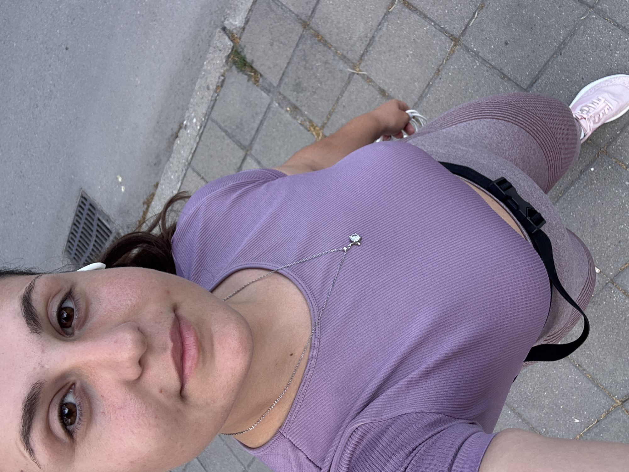 The media file is a portrait of a young woman, taken from a high angle, likely a selfie. She is outdoors, standing on a paved walkway with a gray, gritty texture, bordered by concrete tiles. To the left, a metal grate, possibly a storm drain, is visible. The woman is wearing a lilac-colored, ribbed short-sleeved top and matching ribbed leggings. A black strap with a buckle is visible around her waist. She has on pale pink athletic shoes. A delicate silver chain with a small, clear charm rests on her top. She has light brown eyes, dark eyebrows, and pink lips. She is wearing small white wireless earbuds in her ears. Her hair is dark brown and pulled back. The lighting suggests it is daytime, and the slight shadows indicate a bright but not direct overhead sun. The overall impression is of a person preparing for or in the midst of an outdoor activity, such as jogging or walking. The background suggests an urban or suburban environment. The provided location context is Plovdiv, Bulgaria, which aligns with the paved walkway and urban setting. There is no visible text in the image.