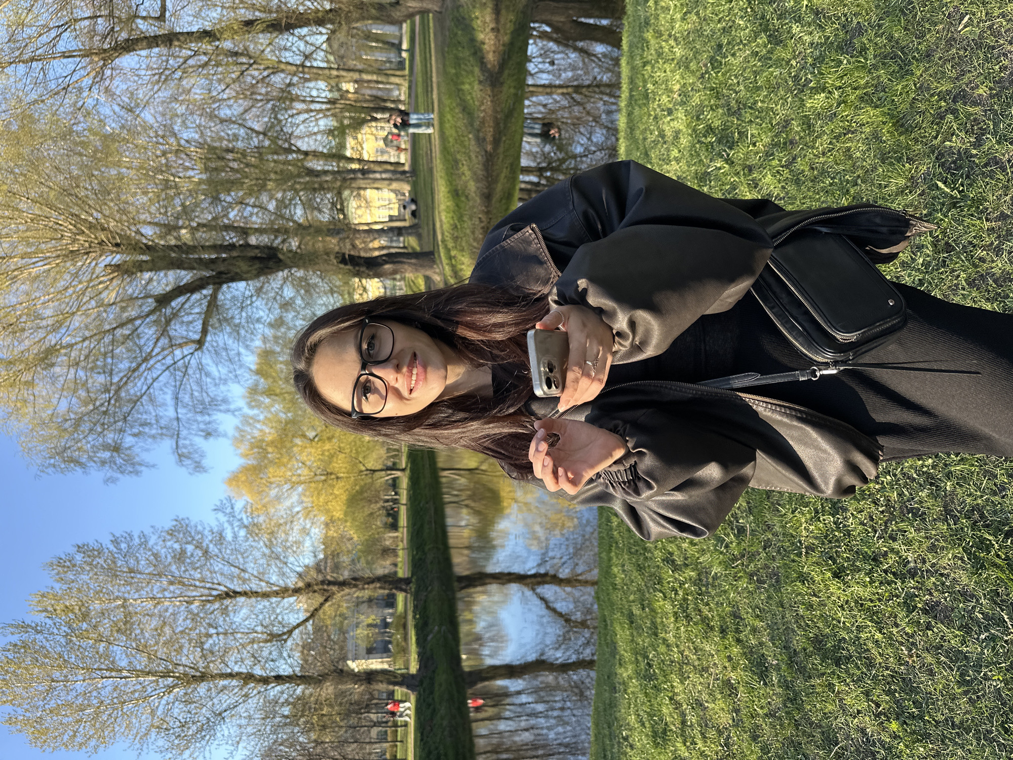A young woman with dark hair and glasses is holding a phone in an outdoor setting. She's wearing a black jacket and has a black purse slung over her shoulder. The background shows a park with trees and a body of water reflecting the sky. This is likely Saint Petersburg, Russia, specifically the area near the Summer Garden and the Neva River. People can be seen in the distance, further indicating the park setting.