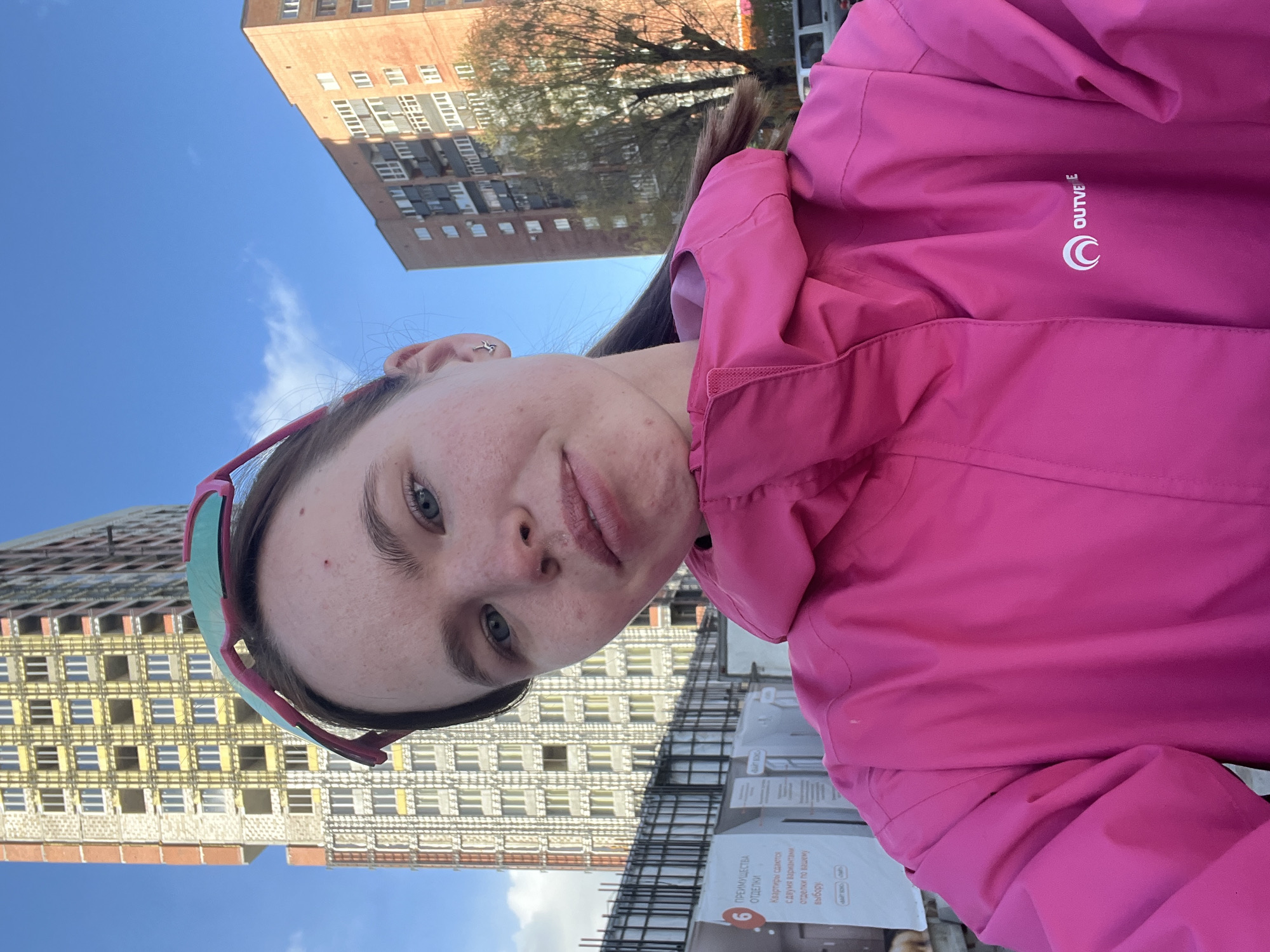 A fair-skinned person with light blue eyes, likely female, is taking a selfie. She wears pink-rimmed sunglasses on her head and a bright pink hooded jacket with a white "OUTVEE" logo on the upper right chest. Her expression is neutral, and some minor skin imperfections are visible.
Behind her, a construction site is partially visible, with rebar and promotional posters hanging on fencing. Two large buildings are in the background. One is an older, light-brown brick building with rows of windows, and the other appears to be a newer construction project, partially unfinished and displaying exposed insulation.
Based on the promotional posters visible, the location is likely a construction site for a new residential building in Perm, Russia.