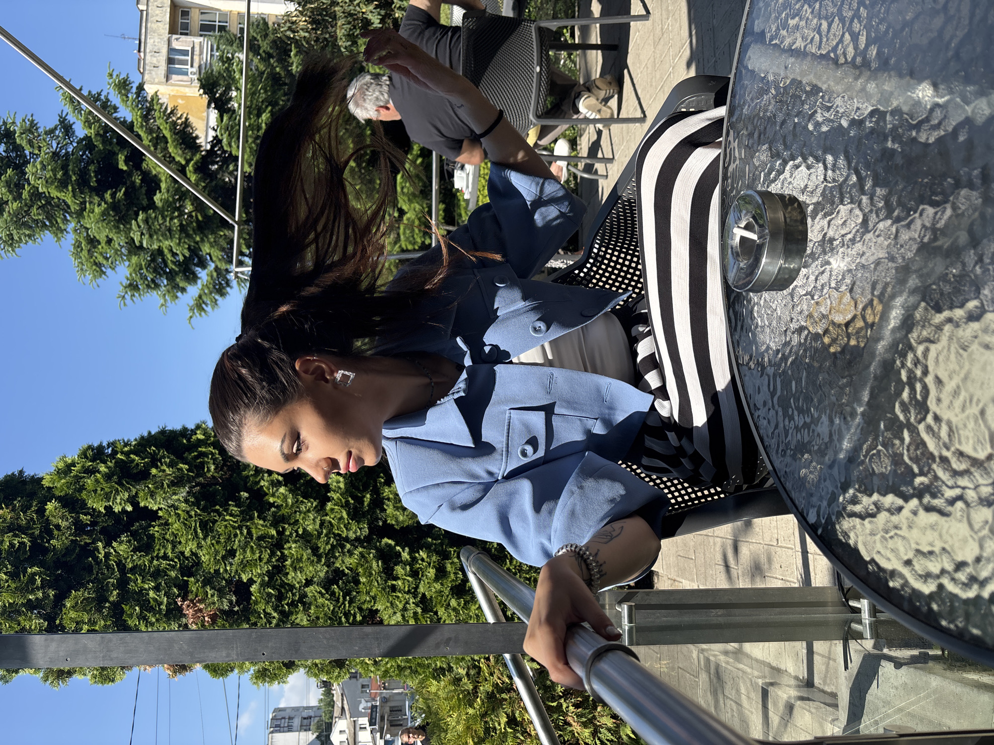This image, rotated 90 degrees clockwise from its natural orientation, captures a sunny outdoor scene, likely a cafe or restaurant patio, in Sofia, Bulgaria.
The central figure is a woman with long, dark hair tied back in a ponytail, which appears dramatically swept to her left, suggesting movement or wind. She is dressed in a light blue blazer or jacket over a lighter top, accessorized with square earrings and a bracelet on her left wrist. Her gaze is directed downwards, conveying a serene or contemplative expression, as she holds onto a metal railing with her right hand.
Beside her, a round, textured glass-topped table is visible, featuring a metallic ashtray. Another chair next to the woman's seat displays a distinctive black and white striped pattern, possibly a cushion or an item of clothing. In the background, a man with gray hair, wearing a dark t-shirt and sandals, is seated at another table, facing away from the camera.
The setting is characterized by lush green trees and bushes, providing a vibrant natural backdrop under a clear blue sky. Parts of several buildings, including an older yellow-toned structure with multiple windows, are discernible behind the foliage, indicative of an urban environment. The bright sunlight casts distinct shadows, underscoring the daytime period. No visible text is present in the image. The overall scene depicts a casual moment of leisure or posing outdoors.