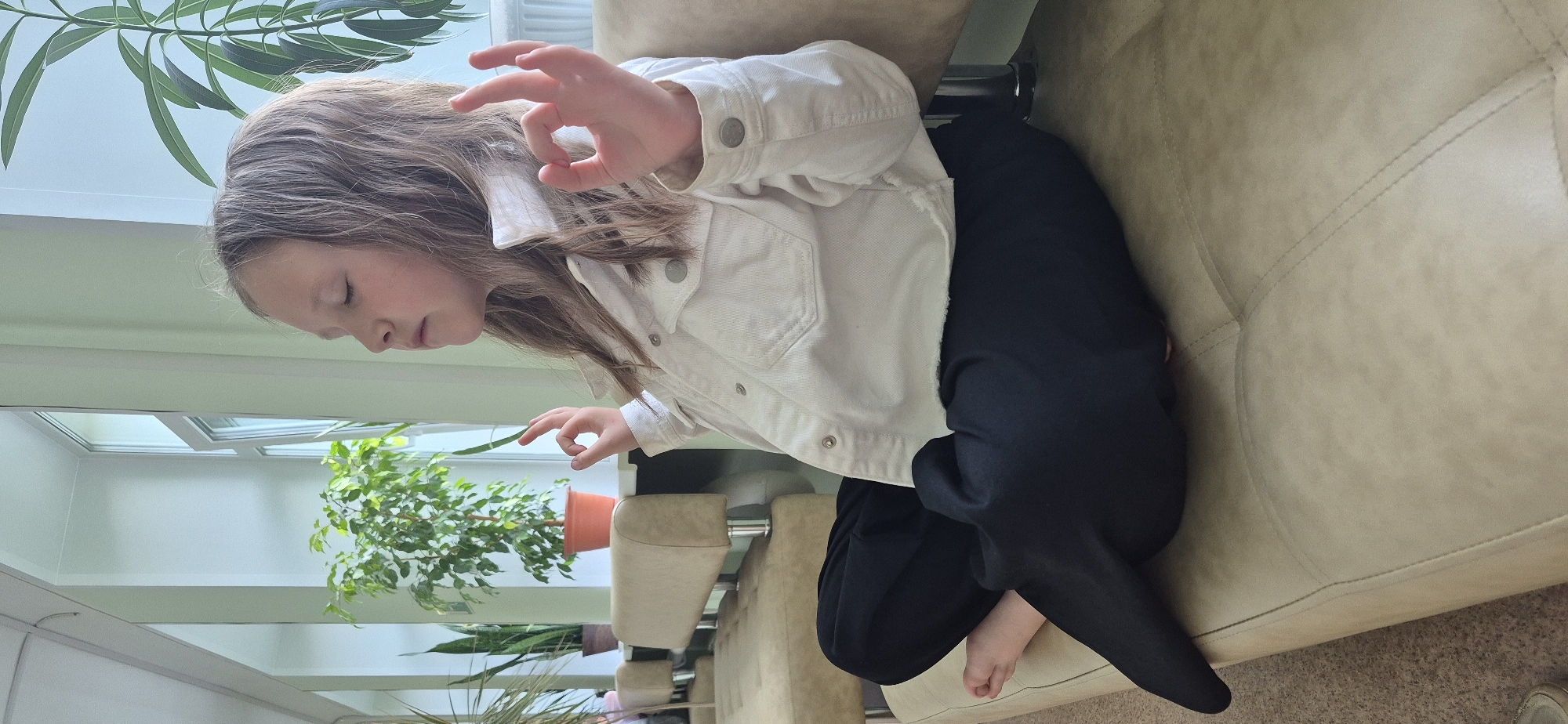 A child with light brown hair, wearing a white denim jacket and dark trousers, is seated on a beige upholstered couch in a room with a skylight overhead. The child has her eyes closed and is holding her right hand up with her thumb and forefinger forming a circle. A potted plant with green leaves is visible in the background, situated near a window. The environment appears to be a well-lit interior space.