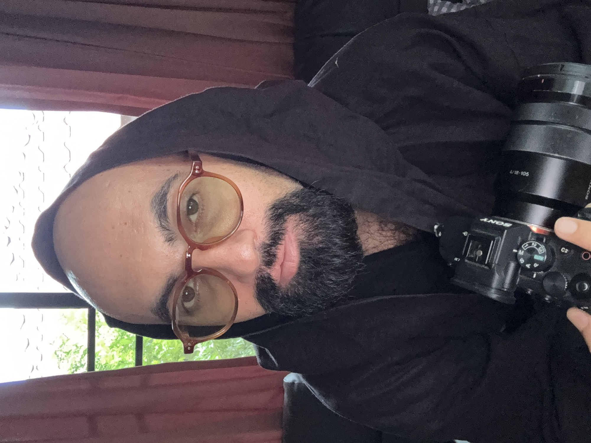 A close-up selfie shows a bald man with glasses and a beard wearing a black hooded jacket. He is holding a black Sony camera with a lens. The man is looking at the camera. He is indoors, and a red curtain is visible behind him. In the background, through a window, green trees are visible. The man's skin tone is light brown, and he has light brown eyes. The camera is pointed at the viewer, and the man's right hand is visible, holding the camera. The lighting is natural, coming from the window and the room.