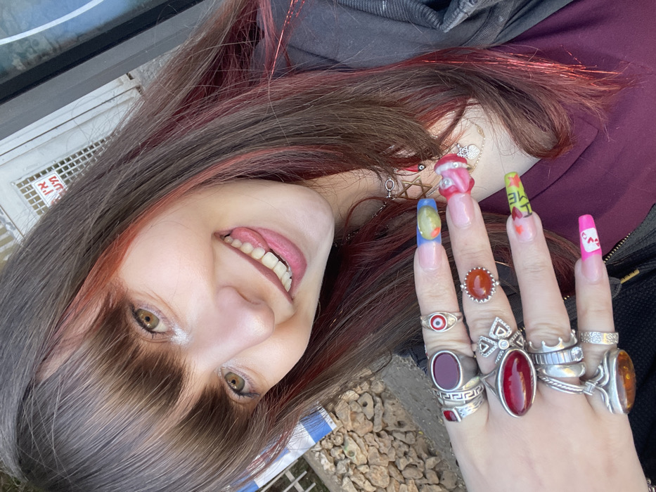 A young adult female is depicted in a close-up, waist-up portrait, smiling broadly with her tongue slightly extended towards the viewer. She has long, dark brown hair with reddish-pink highlights and bangs. Her eyes are light brown/hazel. She wears a dark jacket over a deep red top. Multiple layered necklaces adorn her neck, including one with a silver Star of David pendant, another with a red bead and a small charm, and a delicate chain with a heart pendant. Her left hand is prominent, heavily accessorized with numerous silver rings, many featuring large, polished stones in dark red, orange, and amber hues. One ring displays a red and white evil eye design. Her long, square-tipped fingernails are individually decorated with diverse colors and patterns, including blue, green, yellow, pink, white, red, and black text. The background consists of a textured, light-colored wall or fence on the left, partially obscuring a blue and white striped object. On the right, a darker, reflective surface is visible. Centrally, a light-colored metal gate with a grid pattern features a white sign bearing black Hebrew text: "אין כניסה" (Ein Knisa), meaning "No Entry." The scene is outdoors under natural lighting. This location is in the city of Raanana, Israel.