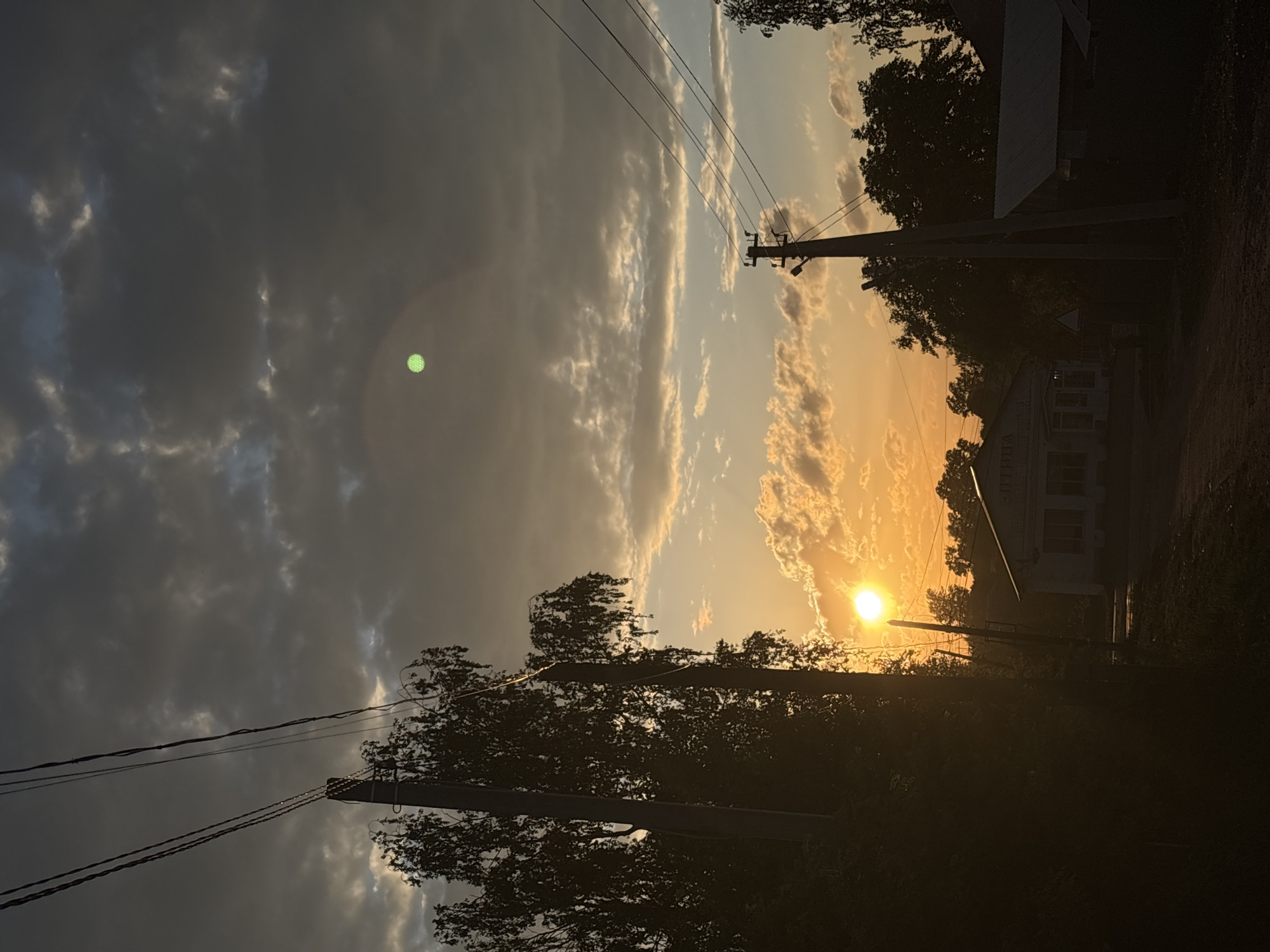 The image captures a sunset in Фоки, Russia. The sky is filled with clouds, some dark gray and others reflecting the golden light of the setting sun. The sun itself is visible on the right side of the frame. Utility poles with power lines stretch across the image, adding a sense of perspective. Silhouetted trees frame the scene at the bottom and along the sides. In the distance, a building is partially visible.