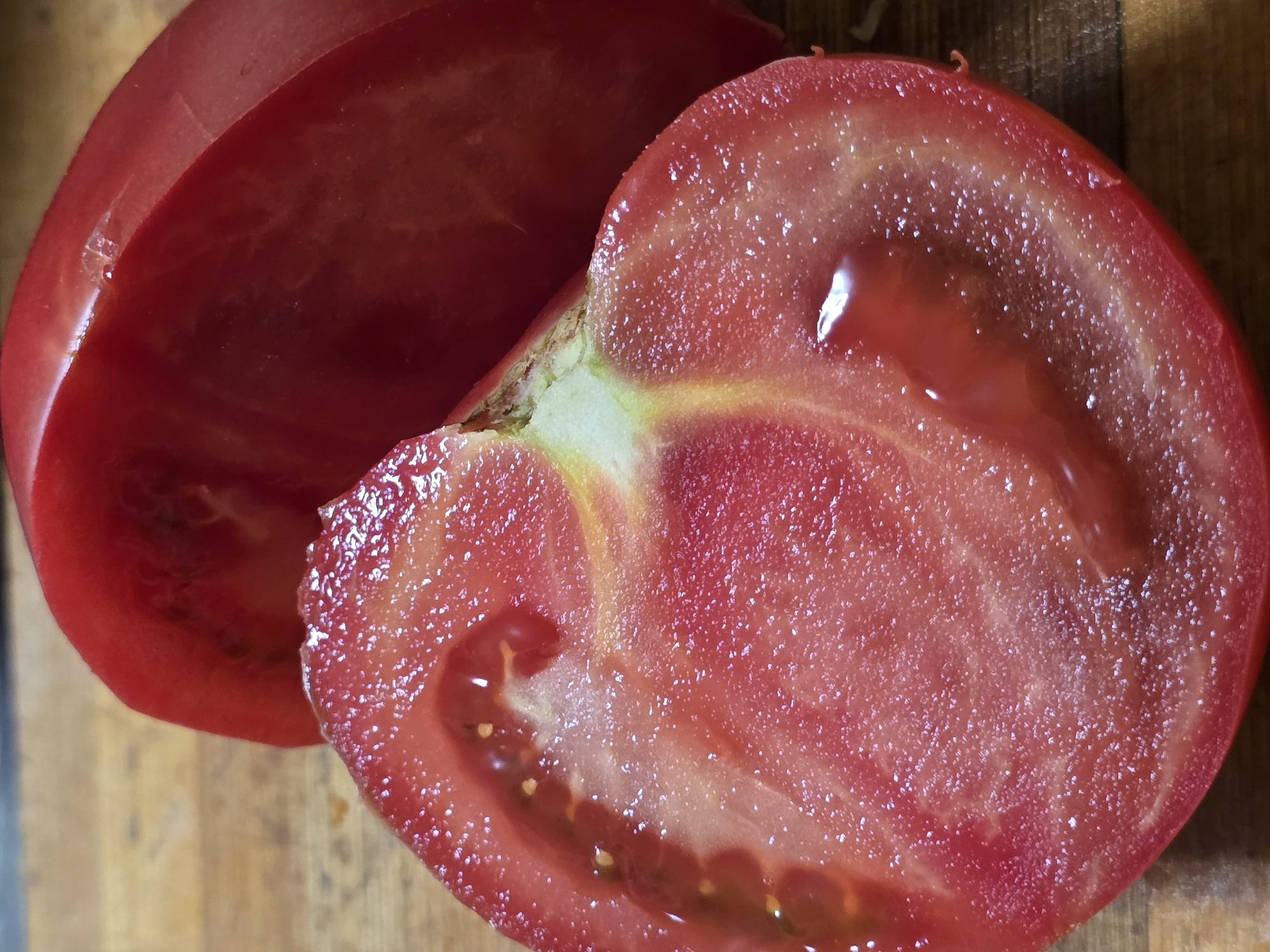 This is a close-up, overhead shot of a sliced tomato on a wooden cutting board. The tomato is bright red with visible seeds and a juicy interior. One half of the tomato is fully visible, showing its internal structure, while the other half is partially obscured by a larger, whole portion of the tomato. The lighting highlights the glossy texture of the tomato flesh. The wooden surface of the cutting board has a visible grain and appears slightly worn. There are no people, animals, or other objects present in the image. The scene does not provide any information that would allow for its localization to Nova Kakhovka, Ukraine, or any other specific city.