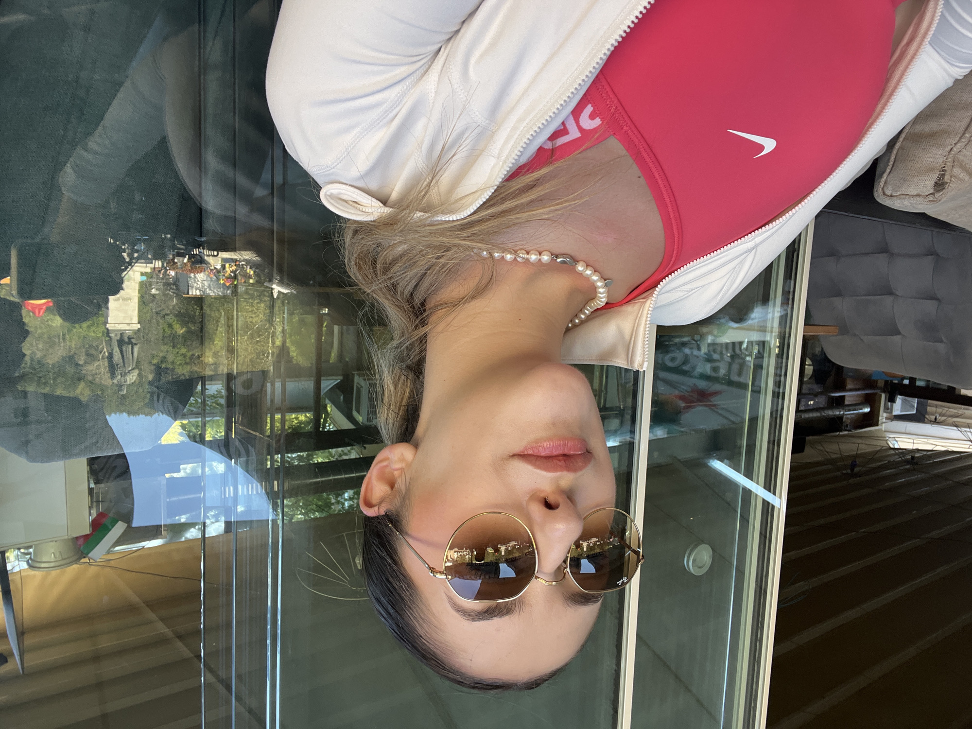 A person is seen from the chest up, with their head facing down. They are wearing a pink sports bra with a white Nike logo and a white jacket. A pearl necklace is visible around their neck. They are wearing round, gold-rimmed sunglasses that reflect a cityscape, possibly a park with trees and buildings. The background shows an interior with glass panes reflecting an outdoor scene. Given the reference to Blagoevgrad, Bulgaria, and the visible reflection, the scene may be from inside a building overlooking a park or cityscape within the city.