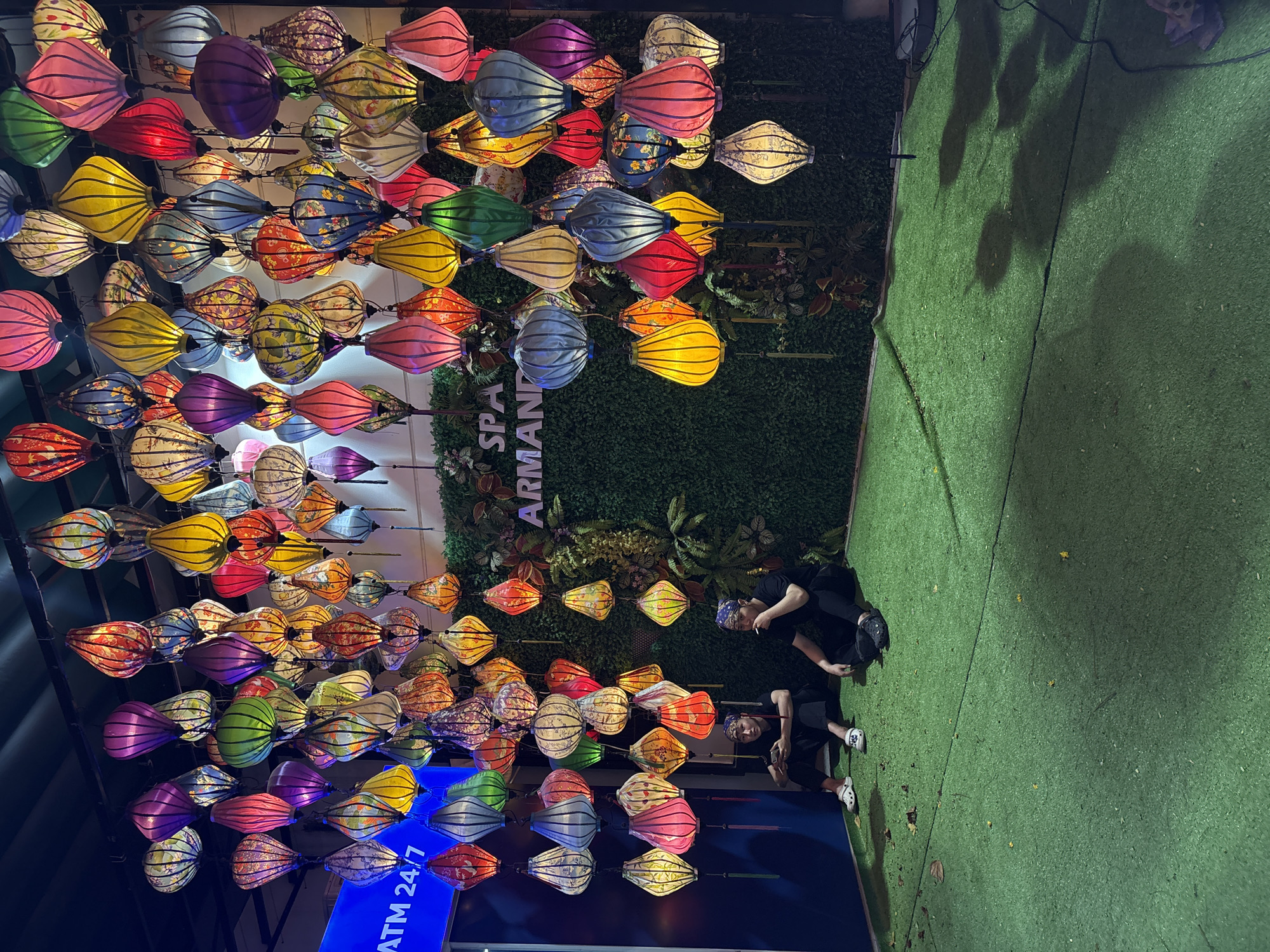 The image presents a vibrant night scene in Đà Nẵng, Vietnam, showcasing a voluminous display of colorful traditional lanterns. Hundreds of illuminated fabric lanterns, primarily in oval and teardrop shapes, hang suspended in dense rows, creating a glowing canopy overhead. These lanterns feature a diverse array of patterns, including intricate floral and geometric designs, and are rendered in a rich spectrum of hues such as pink, purple, yellow, red, green, blue, and white.
Behind the extensive lantern arrangement, a vertical green wall adorned with artificial foliage forms a contrasting backdrop. White letters partially visible on this wall spell out "SPA ARMAN D." To the right of the lantern display, the ground is covered with bright green artificial turf. Two individuals, appearing to be young men, are casually seated on this turf. Both are dressed in dark clothing; one wears a blue patterned bandana and is holding a small object, while the other is looking downwards. Their relaxed postures suggest a moment of rest or casual observation.
The setting appears to be an outdoor or semi-outdoor commercial or recreational area, possibly a night market or a decorated plaza. The bright illumination emanating from the lanterns clearly indicates it is nighttime. A blue sign with white text, partially visible at the bottom left, reads "ATM 24/7." The overall atmosphere conveyed by the glowing lanterns is festive and warm, characteristic of cultural displays in Southeast Asia.