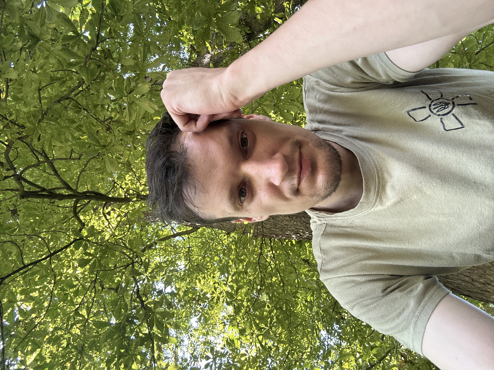 A man is taking a selfie outdoors, leaning against a tree. He is wearing a light green t-shirt with a symbol on the left chest area. His right hand is resting on his forehead. The background consists of dense green foliage and tree trunks, suggesting a wooded area. The scene is illuminated by natural sunlight, creating dappled light effects on the leaves. The location is identified as Makiv, Ukraine.