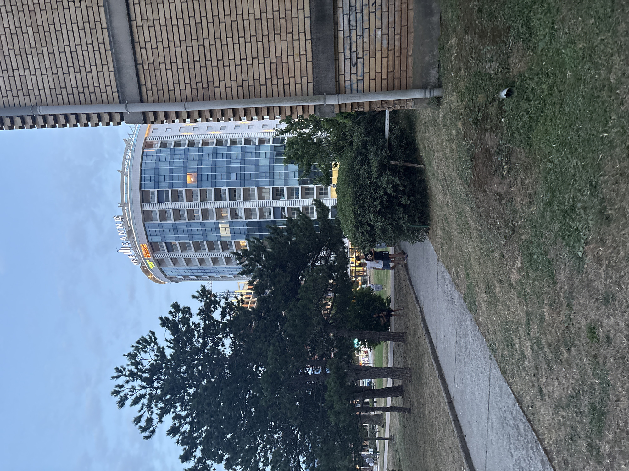 The image shows a modern, curved building with a glass facade, likely an office building or hotel, in Budva, Montenegro. The building has many windows, and some appear to be lit from within, suggesting it's either late afternoon or evening. A neon sign on the building is partially visible, displaying some text. In the foreground, there are several tall pine trees and a grassy area with a paved walkway. Two people are walking on the walkway, appearing to be a couple, with one possibly holding a dog's leash. The sky is clear and blue, indicating fair weather. The overall scene depicts an urban environment with green spaces, common in a coastal city like Budva.