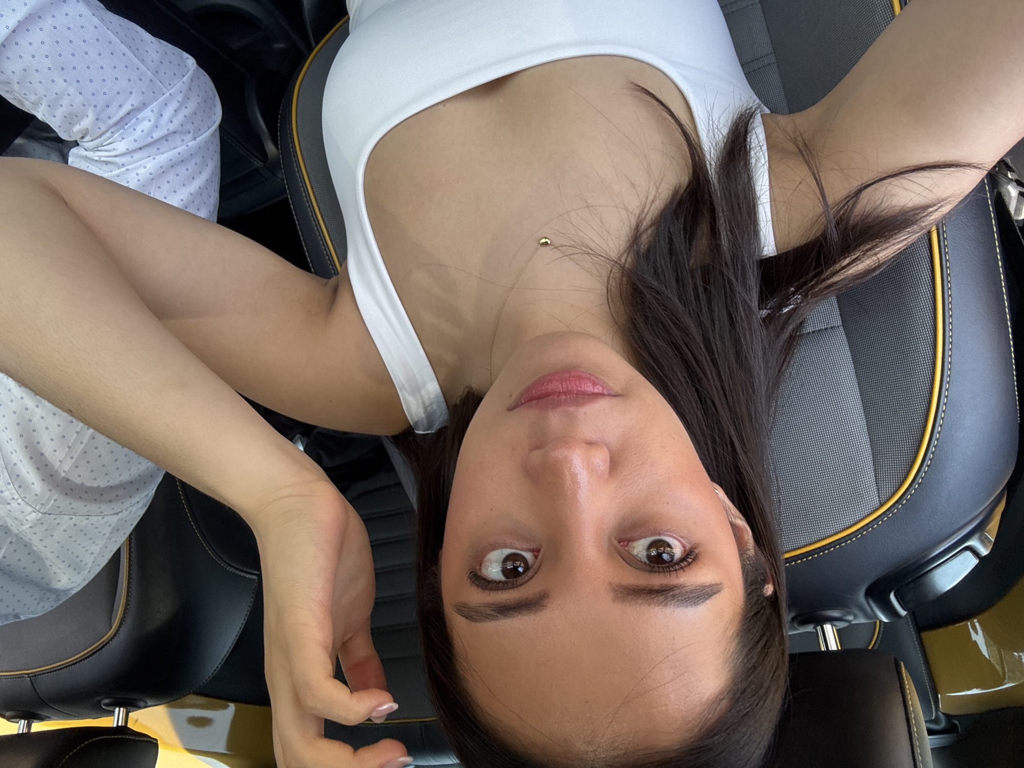 A woman with dark hair and brown eyes is lying on her back in the front passenger seat of a vehicle. She is wearing a white tank top and a delicate gold necklace. Her skin tone appears light. She is looking directly at the camera with a somewhat wide-eyed expression. To her left, a person's arm wearing a light blue patterned shirt is visible. The car seats are black with yellow piping and gray inserts. The vehicle interior has a golden hue. The photo was likely taken from directly above her. The location is assumed to be in Puebla, Mexico, based on contextual clues not visible within the image.