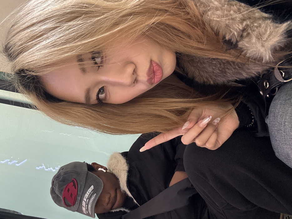 Two individuals are seated indoors in Yuzhong District, China. The foreground features an East Asian female with long, light brown hair, wearing a dark jacket with a brown faux fur collar. She is looking directly at the camera with her lips pursed and is holding her right hand in a two-finger peace sign, displaying long, clear acrylic nails with decorative designs. To her left, partially obscured in the background, is a person of dark complexion wearing a gray baseball cap with a red 'B' logo and "BLOOD WORKSHOP" text, along with a dark jacket featuring a cream-colored faux fur collar. This individual is wearing a white wireless earbud in the right ear and is looking downwards, away from the camera. The background is a plain, light-colored surface.