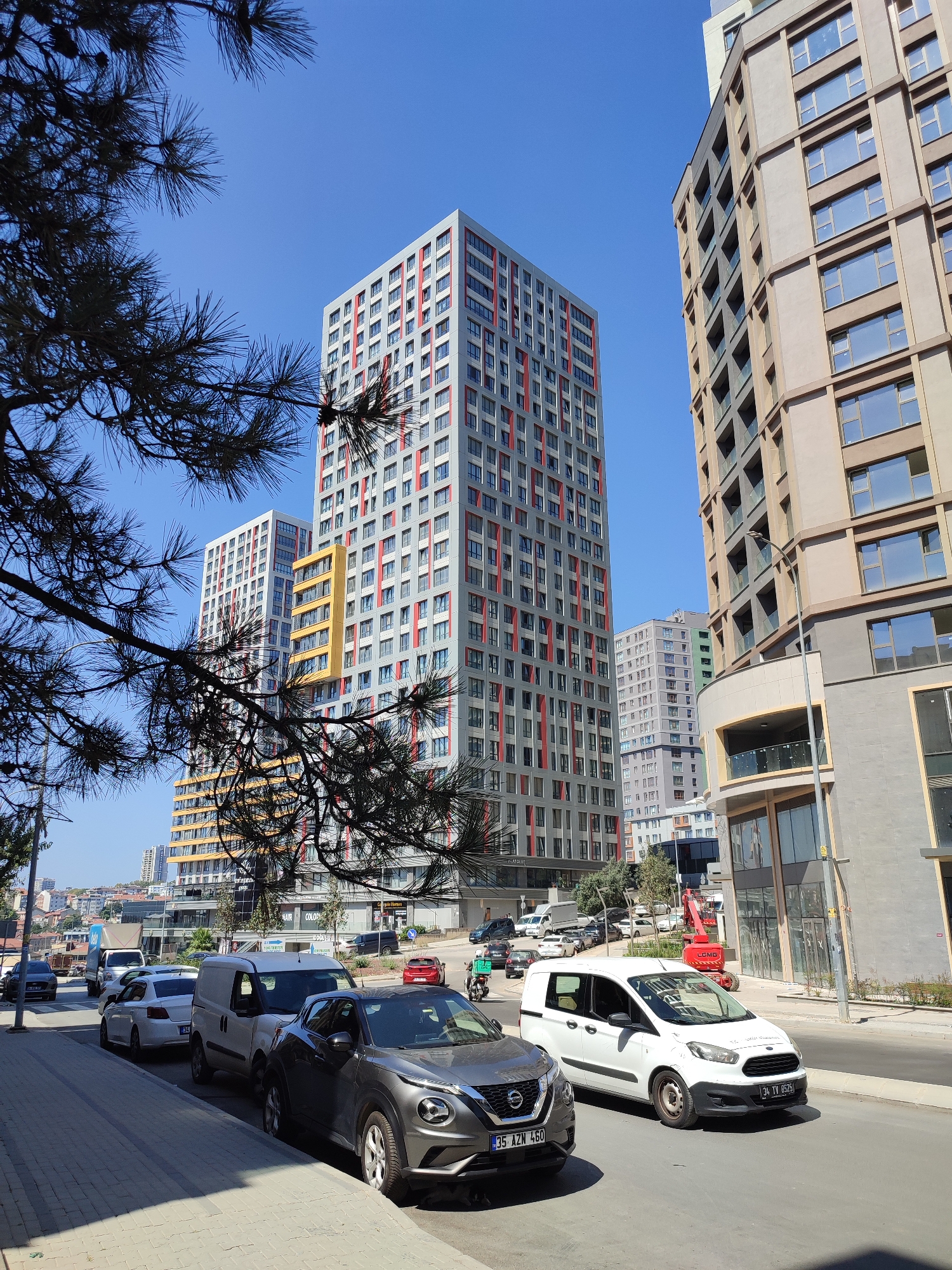 The image shows a street scene in Kadıköy, Turkey, with several modern apartment buildings under a clear blue sky. In the foreground, a gray Nissan Juke is parked on the right side of the street, and a white Ford Transit van is parked next to it. Further down the street, a silver sedan is visible, followed by a white delivery truck and other vehicles. The buildings are tall and have contemporary designs with many windows. A red construction crane is positioned near one of the buildings. The street is paved, and there is a sidewalk with a brick pattern. Pine tree branches are visible in the upper left corner of the frame, casting shadows. The overall impression is of an urban environment on a sunny day.