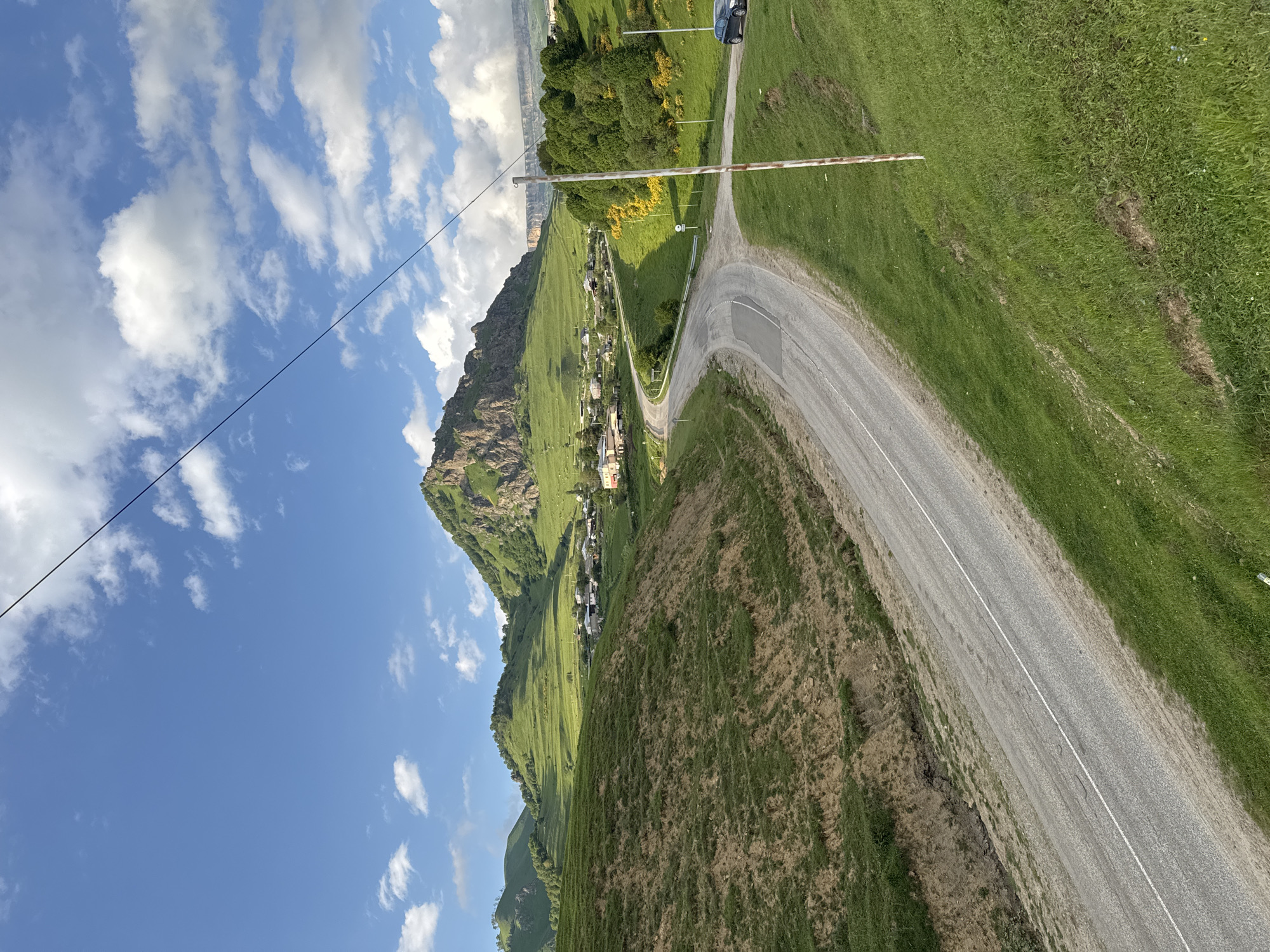 This image captures a scenic view of a winding road traversing a lush, green mountainside in Nizhnyaya Mara, Russia. The scene is bathed in the bright sunlight of what appears to be a clear day, with scattered white clouds dotting the vibrant blue sky. A power line runs diagonally across the upper left portion of the frame, adding a man-made element to the natural landscape.
The road, paved and marked with a white line, curves gracefully around the mountain. On the right side of the road, a verdant, grassy slope dominates the foreground, with patches of dry earth and some erosion visible. A red and white barrier pole is positioned across the road, suggesting a checkpoint or an entrance.
Further up the mountainside, nestled amongst the green slopes, a small village or settlement can be seen. Houses with red roofs are clustered together, giving a sense of community within the vast natural setting. The mountain itself rises dramatically behind the village, its rocky facade partially covered in greenery.
To the left of the road and mountain, the sky takes center stage, with fluffy white clouds creating patterns against the deep blue expanse. A black car is partially visible in the upper right corner, driving on the road. The overall impression is one of natural beauty, tranquility, and a remote mountain community. The time of day is likely midday, given the bright sunlight and clear sky.