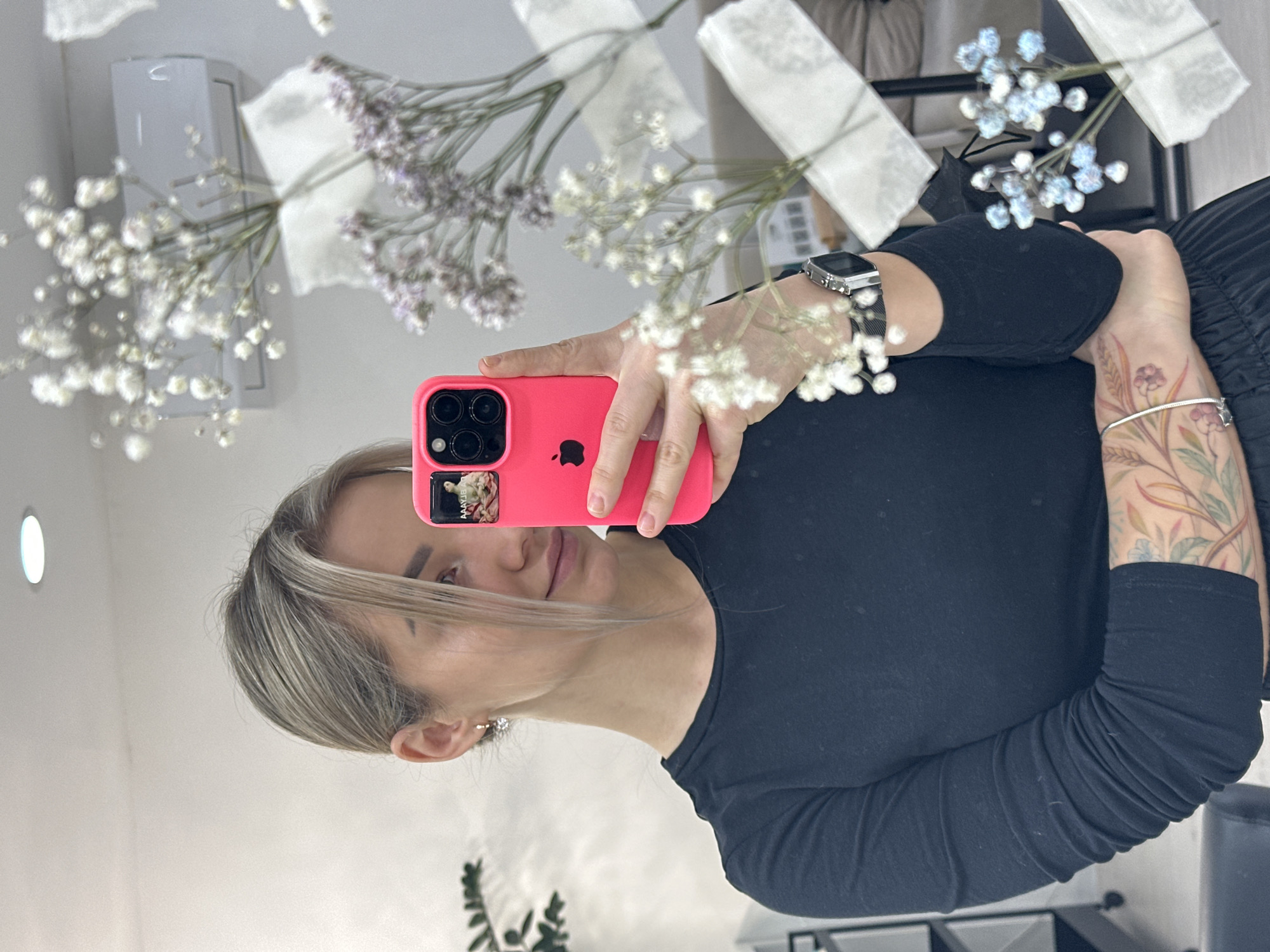 The image shows a woman taking a selfie in front of a mirror. She is holding a pink smartphone, obscuring part of her face. She has blonde hair and a dark long-sleeved shirt. There is a tattoo on her left arm. The background shows a wall with flowers and what seems to be a decorative item or appliance. Based on the style and decor elements, the setting might be a salon or similar beauty-related space. There are no direct visual cues to confirm it's in Budva, Montenegro, but the environment suggests a contemporary, possibly urban location.